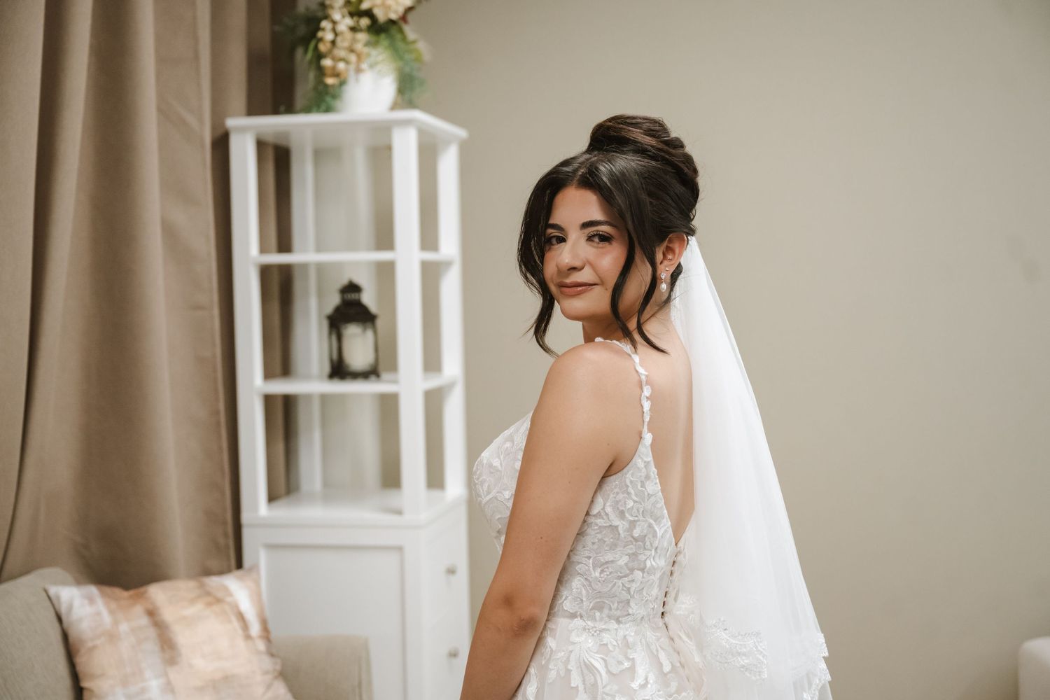 Bride in a lace wedding dress and veil poses elegantly indoors, glancing over her shoulder with a soft smile.