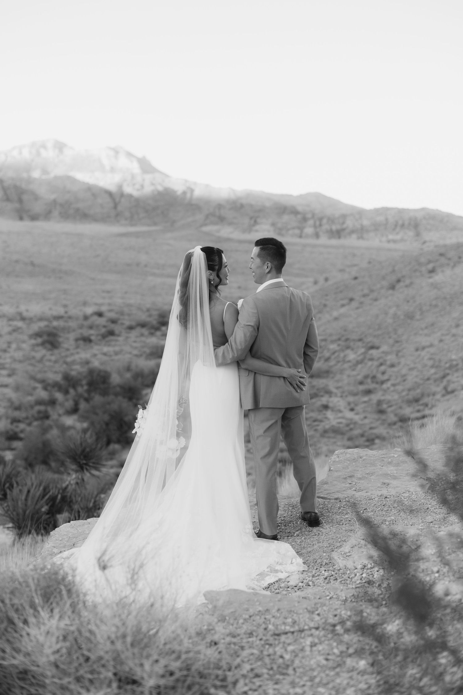 Black and white photo of a bride and groom embracing on a desert path with mountains in the background.