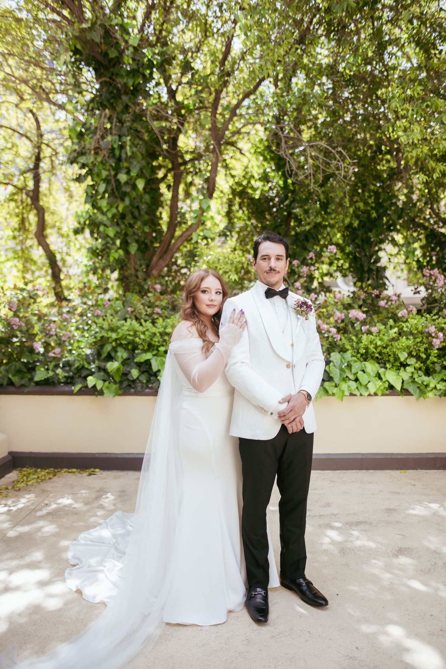 A couple in wedding attire poses together outdoors in a garden setting with lush greenery in the background.