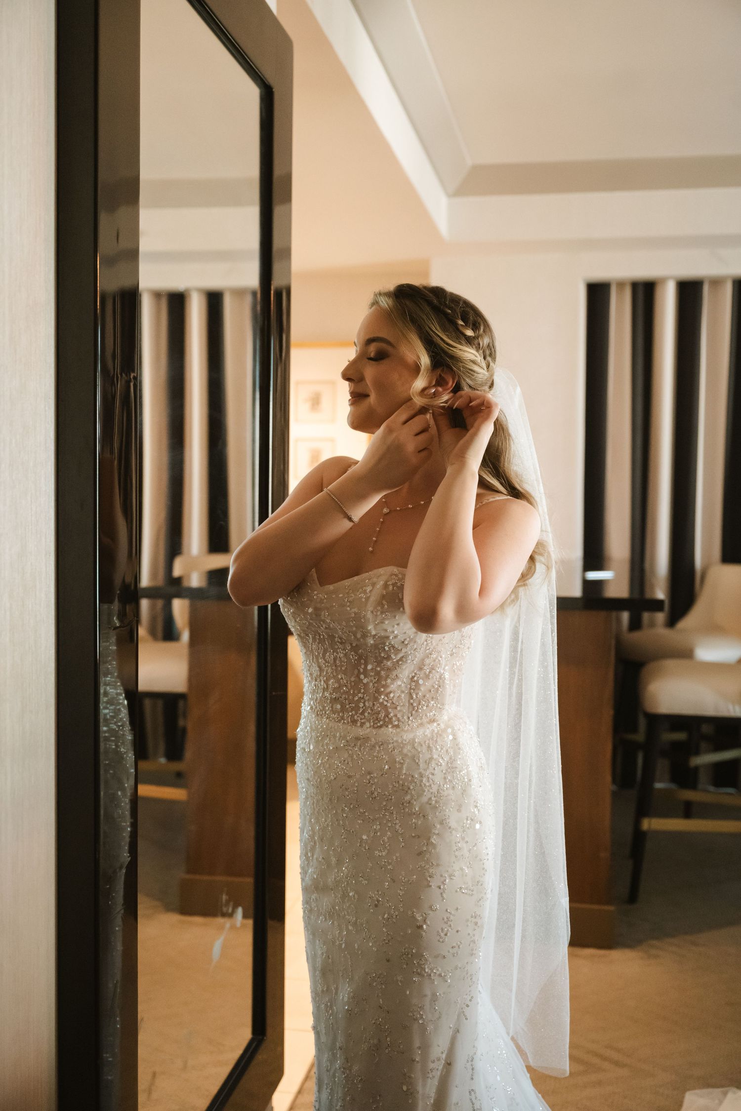 A bride adjusts her earrings while wearing a fitted lace wedding gown and veil.