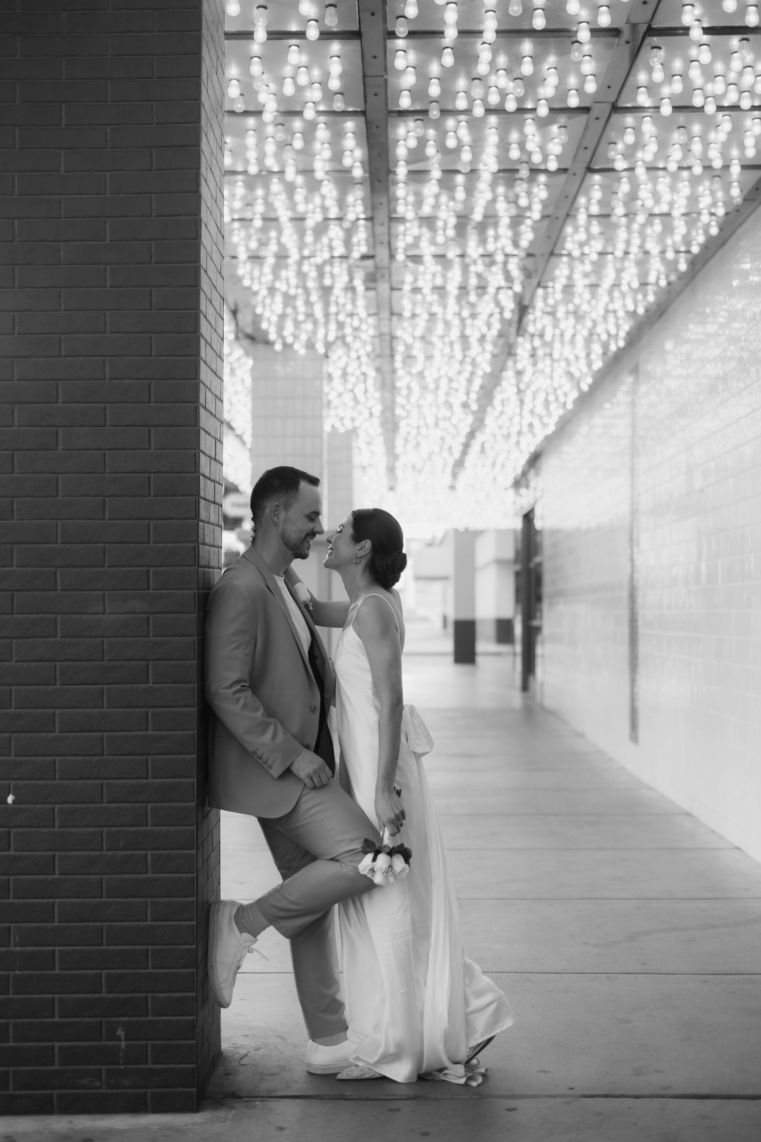 A romantic couple poses in a modern hallway illuminated by twinkling string lights overhead creating a magical atmosphere.