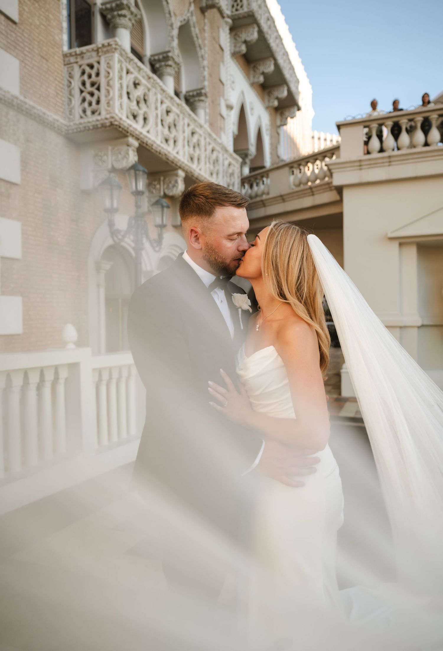 A bride and groom share a romantic kiss outdoors, her flowing veil billowing around them near an elegant building.