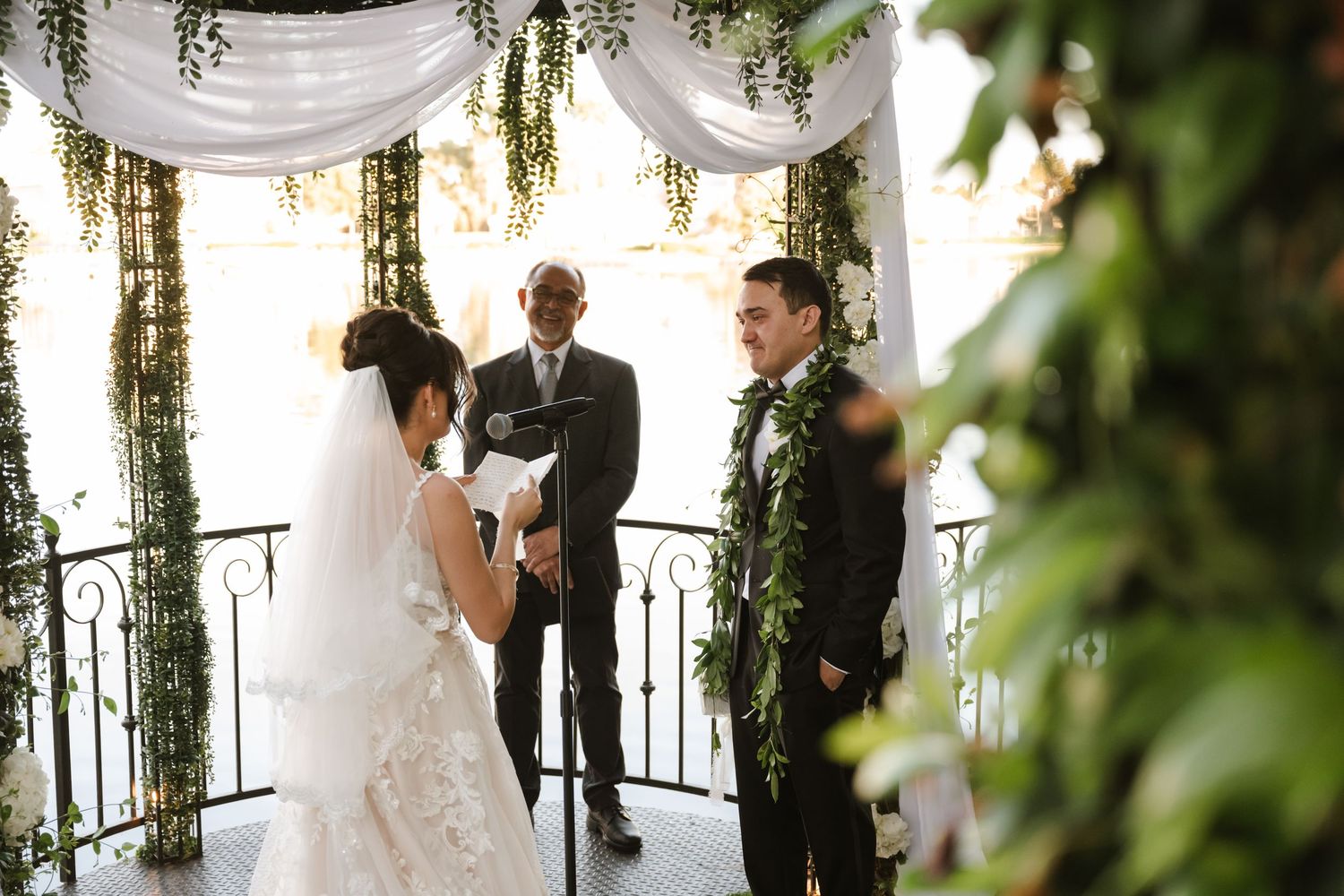 Bride and groom exchange vows before officiant under draped floral arch at outdoor wedding.