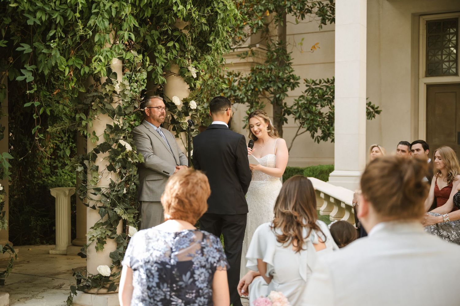 Outdoor wedding ceremony with bride and groom at the altar, surrounded by lush ivy-covered columns and seated guests.