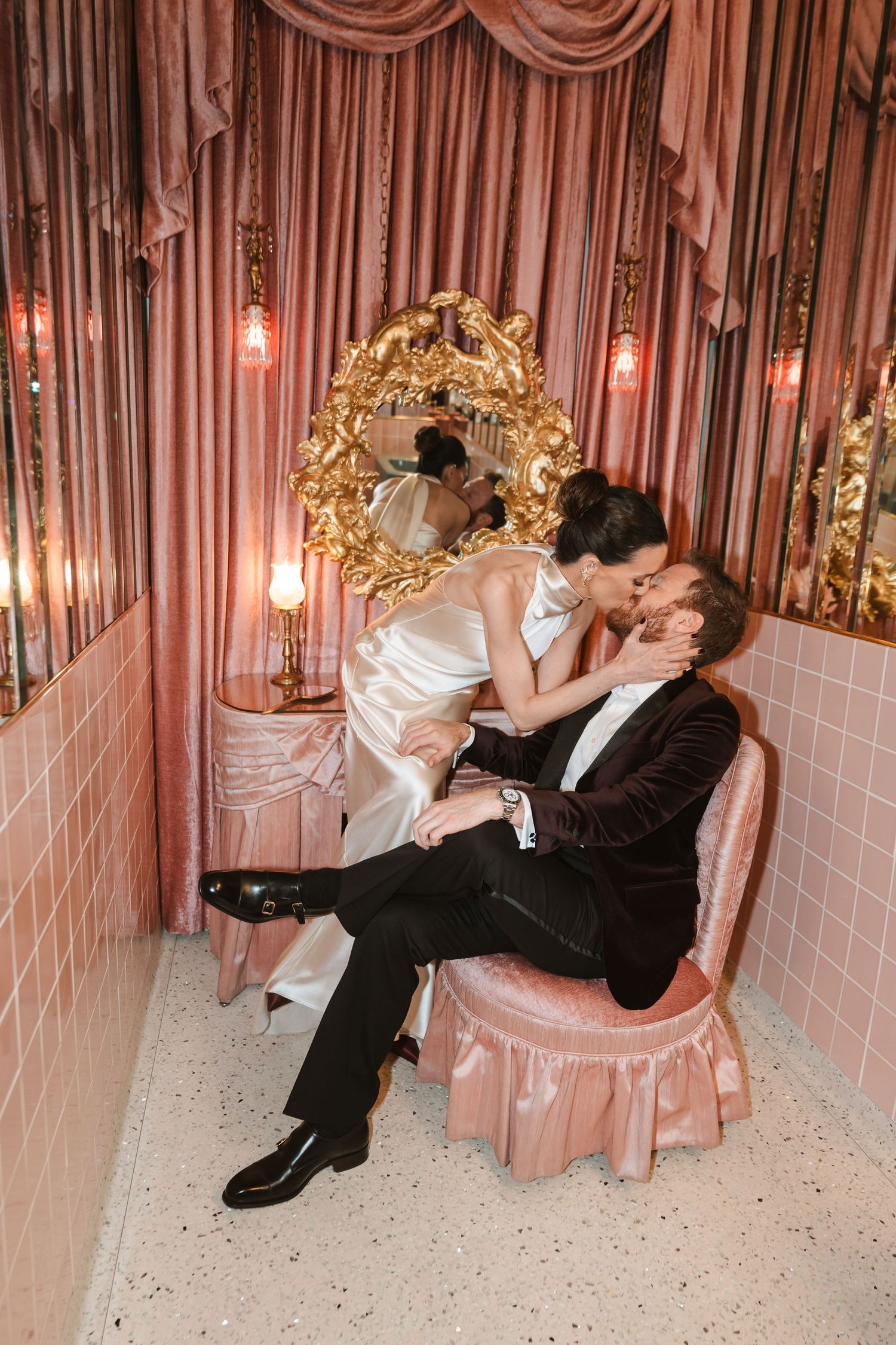 Romantic couple in vintage-inspired setting with pink drapes and ornate gold mirrors sharing intimate moment on pink chair.