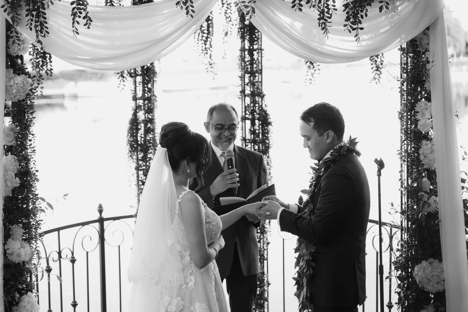 Black and white wedding ceremony photo of bride and groom exchanging vows with officiant under floral arch.