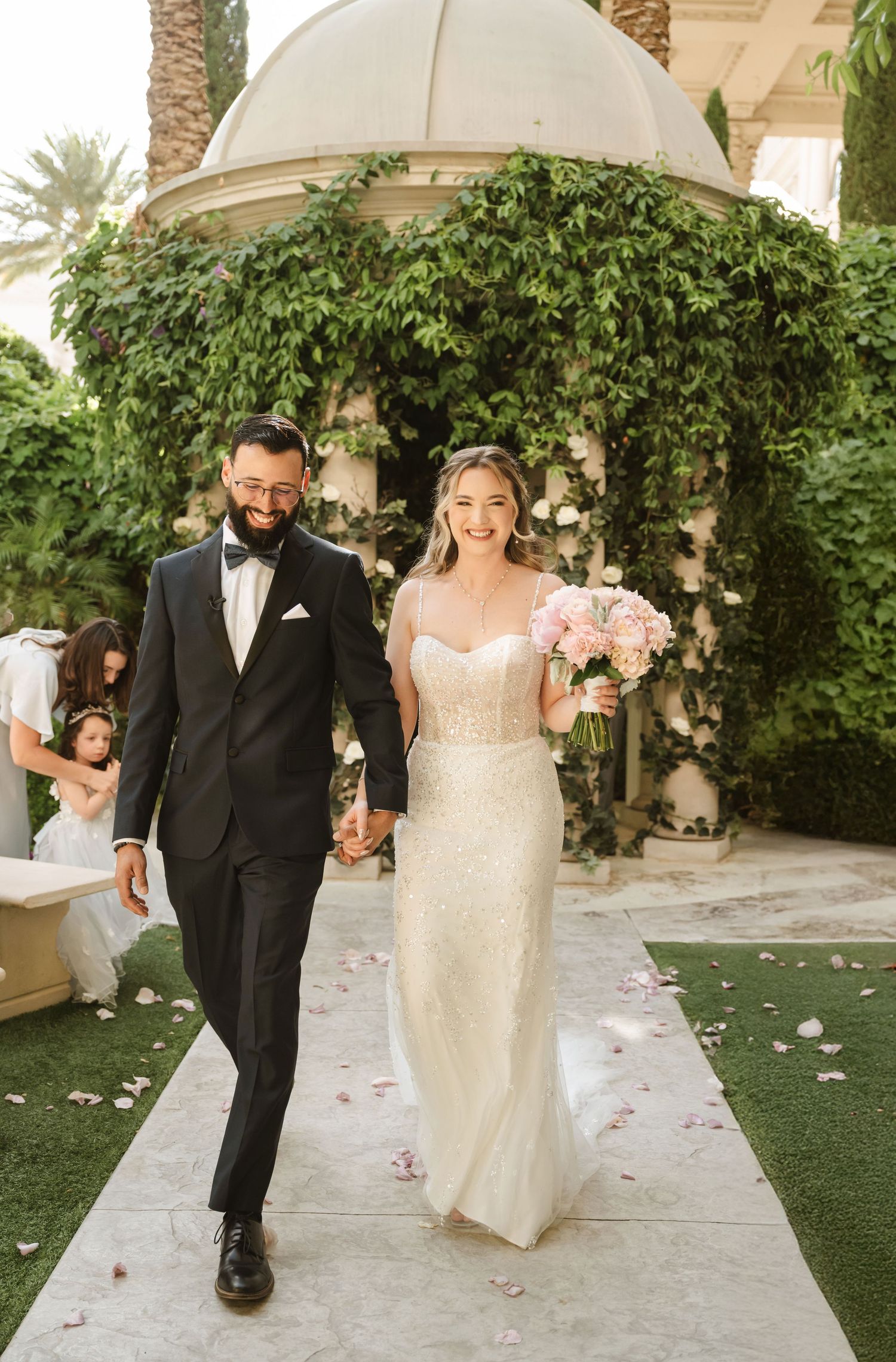 A couple walks down a garden path under a domed gazebo covered in greenery.
