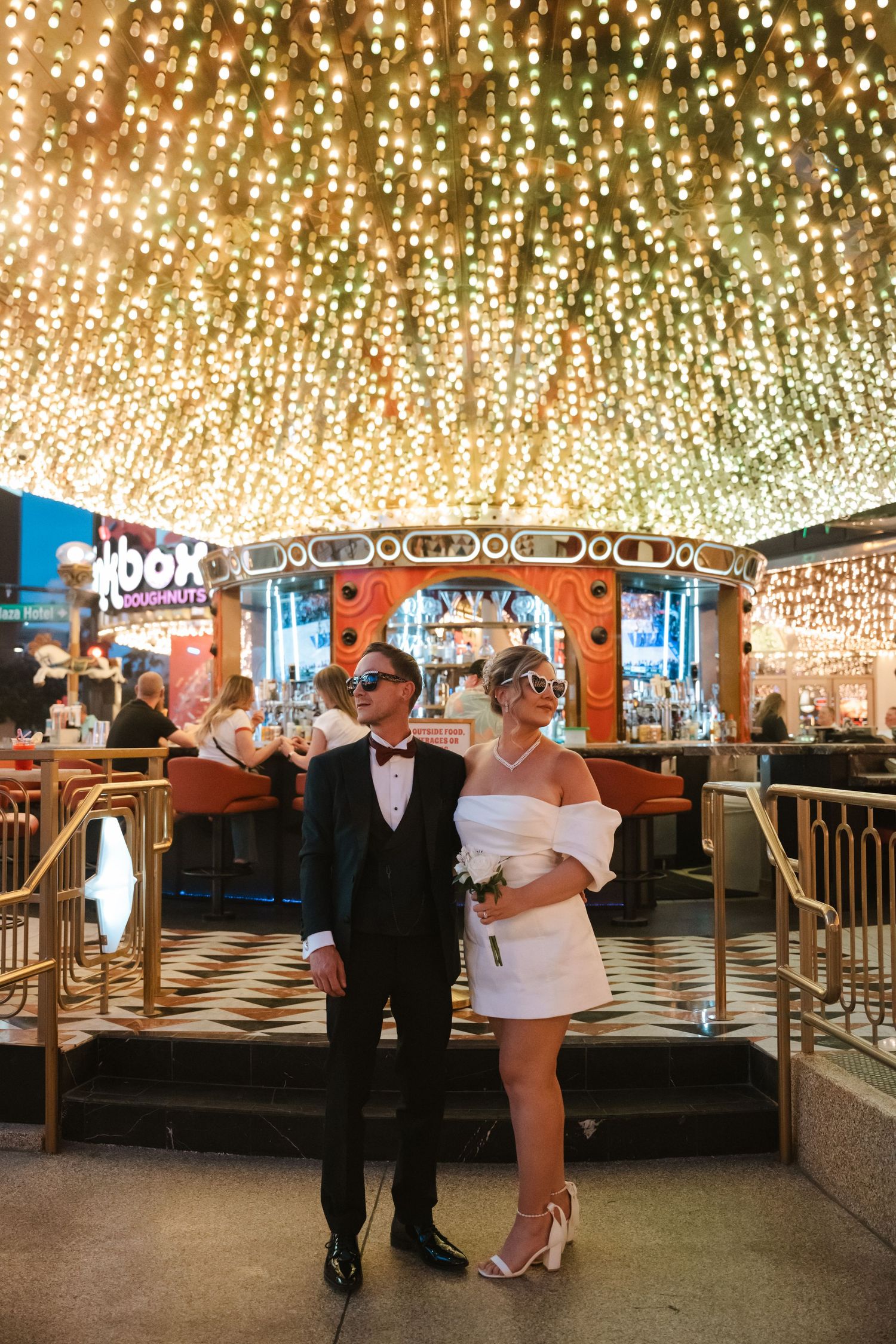 Couple in wedding attire wearing masquerade masks pose under sparkling lights at a Las Vegas venue.