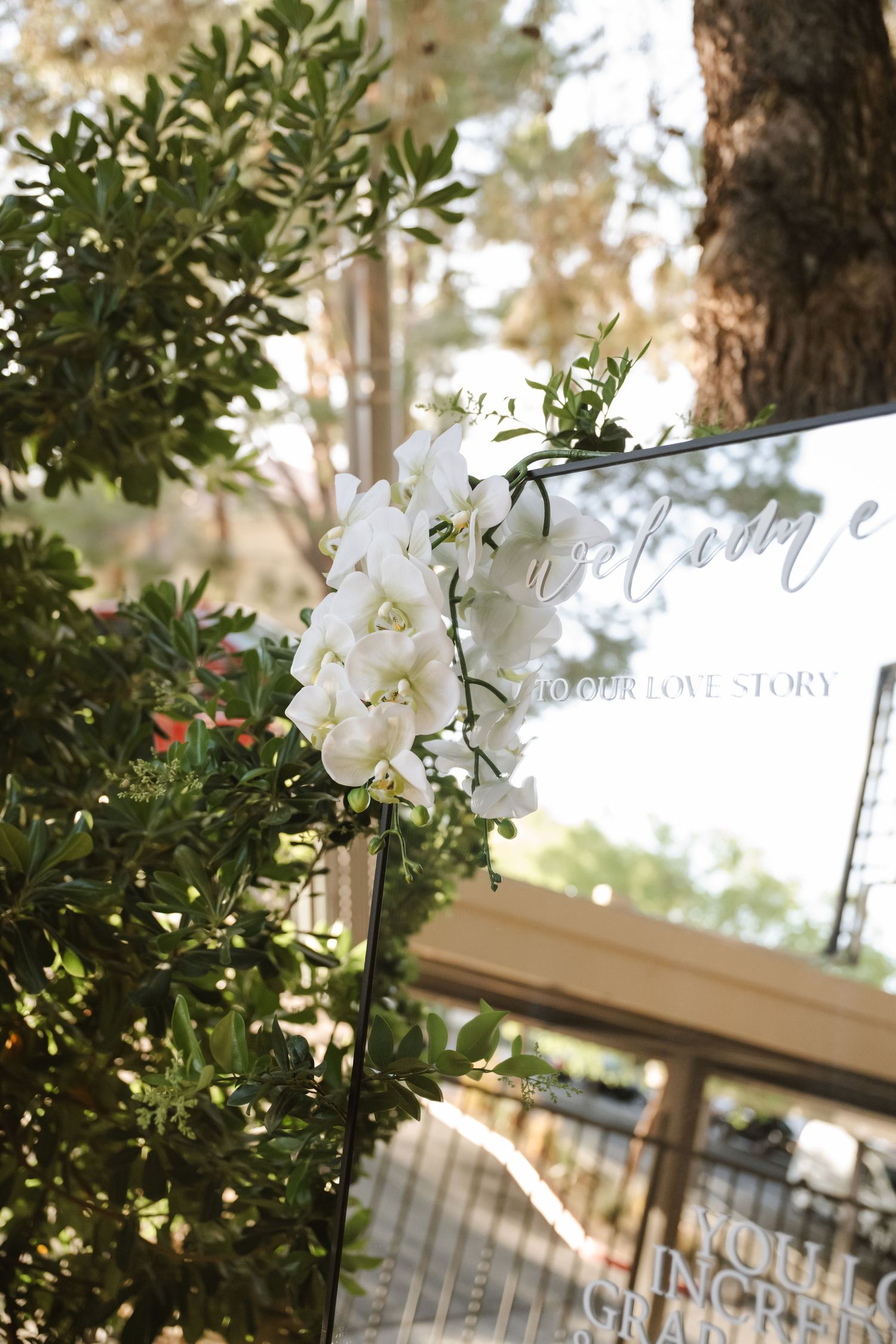 White orchid floral arrangement adorning a wedding welcome mirror sign surrounded by lush green foliage outdoors.