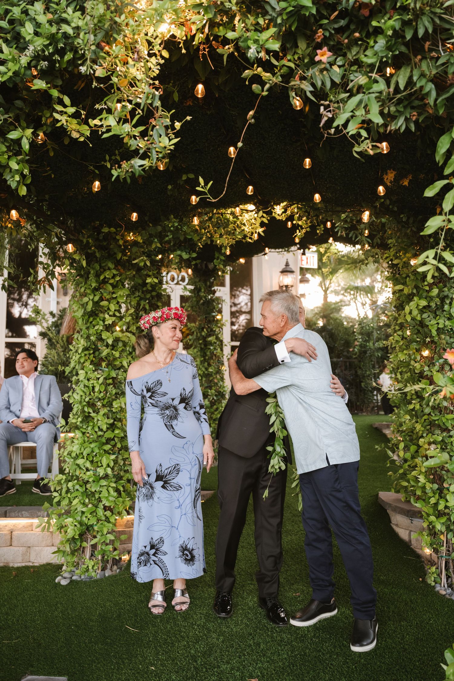 Guests embrace and celebrate under string lights and lush greenery at an outdoor evening wedding reception.
