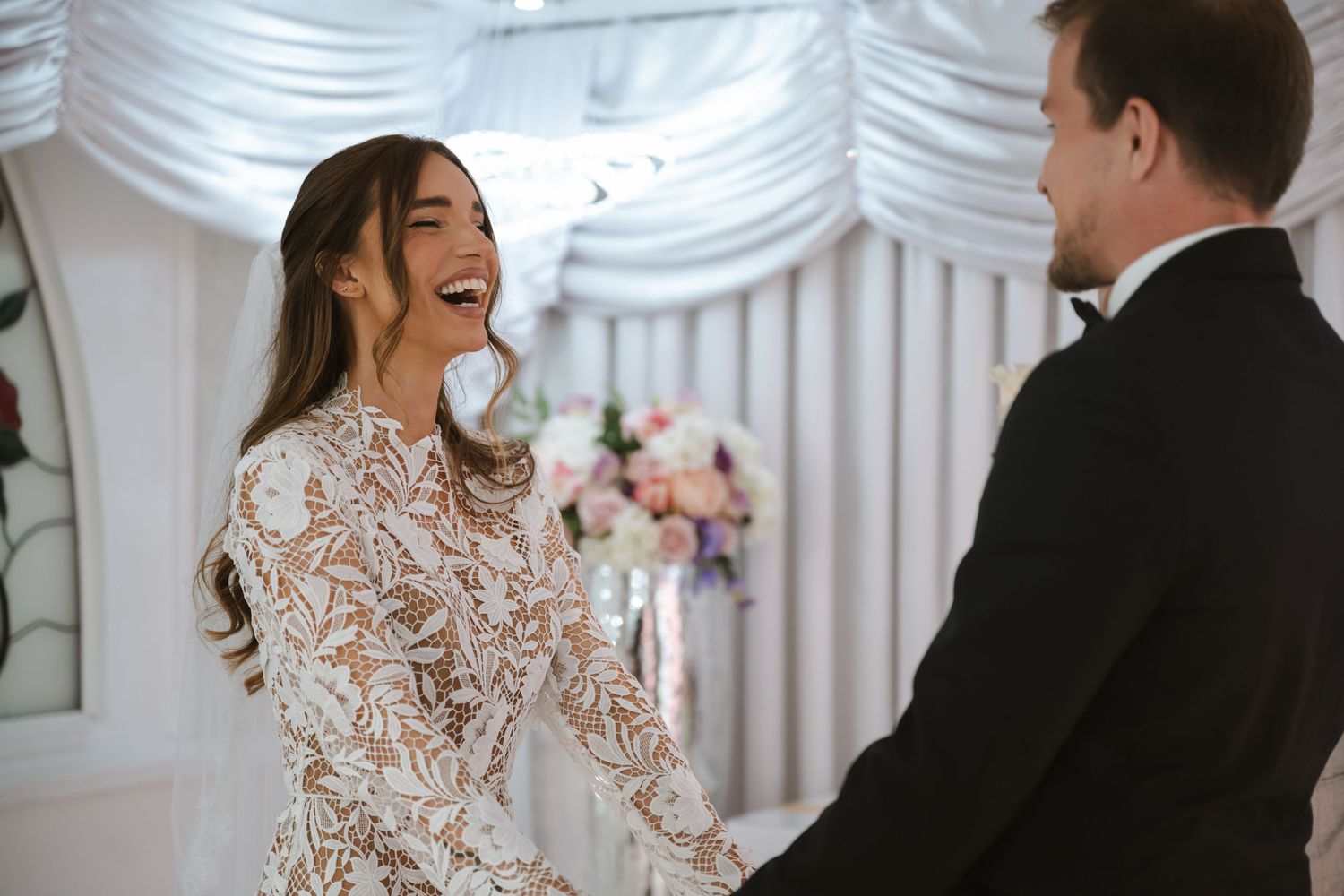 Bride in lace long-sleeve gown laughing joyfully with groom in black tuxedo during wedding ceremony.