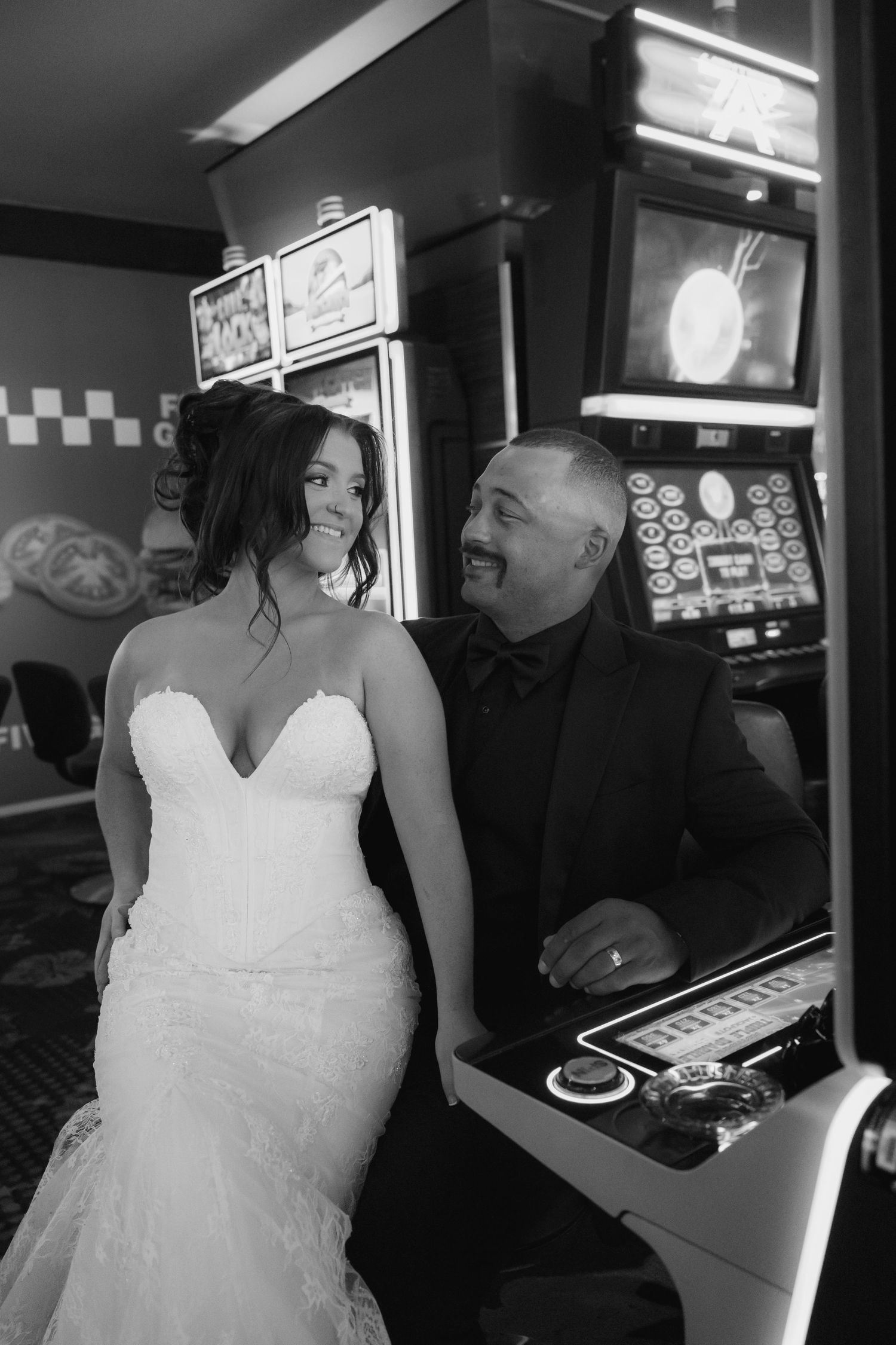 Black and white photo of a bride in a lace gown smiling at a groom in a dark suit seated at a casino slot machine.