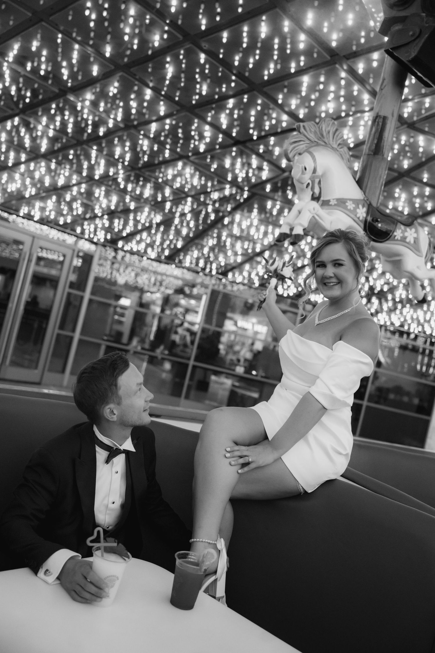 Black and white photo of an elegant couple at a carousel bar, woman in white dress seated on ledge, man in tuxedo beside her.