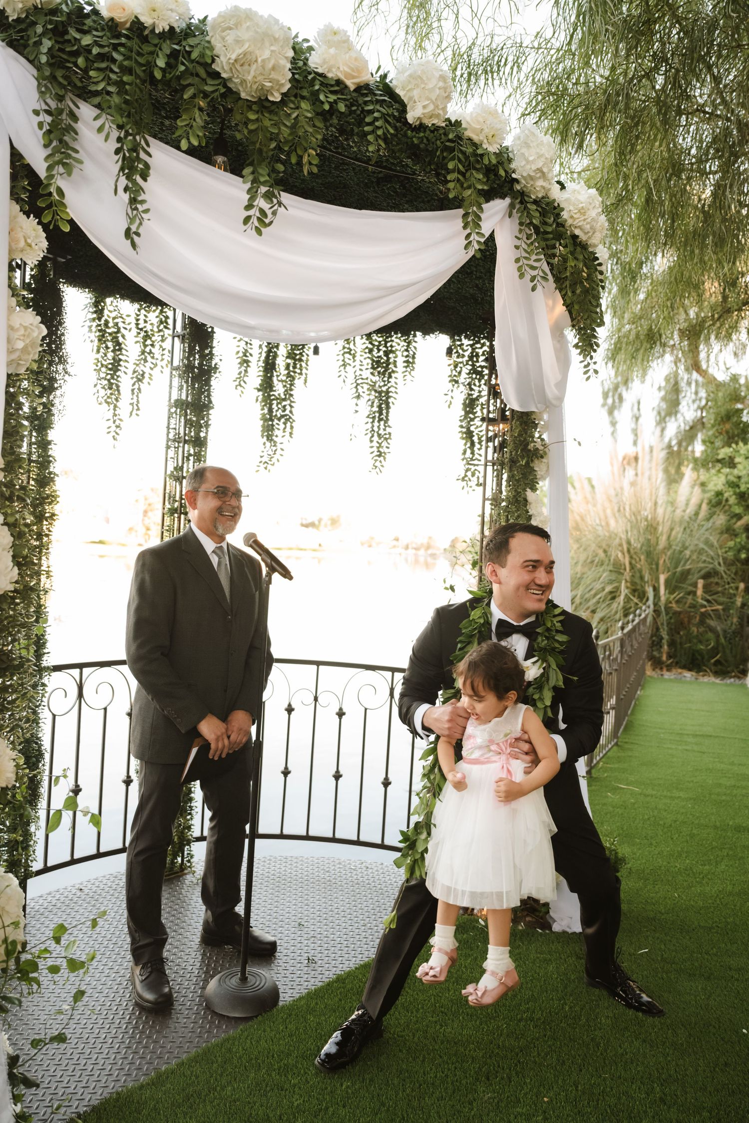 Groom smiles with flower girl under floral arch with officiant at waterfront wedding ceremony.