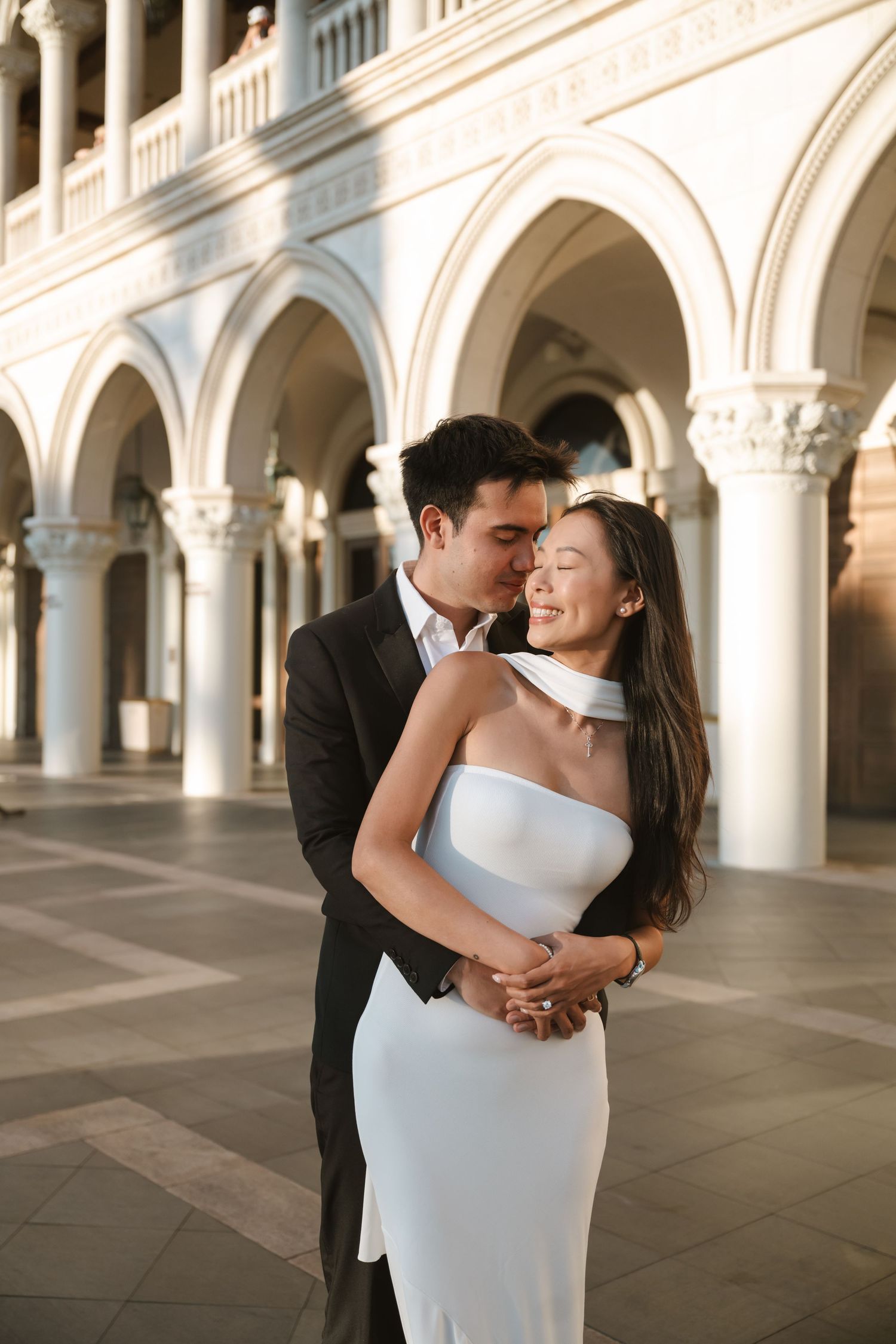 Couple embracing in elegant white dress and suit under ornate arched columns in romantic wedding photo.