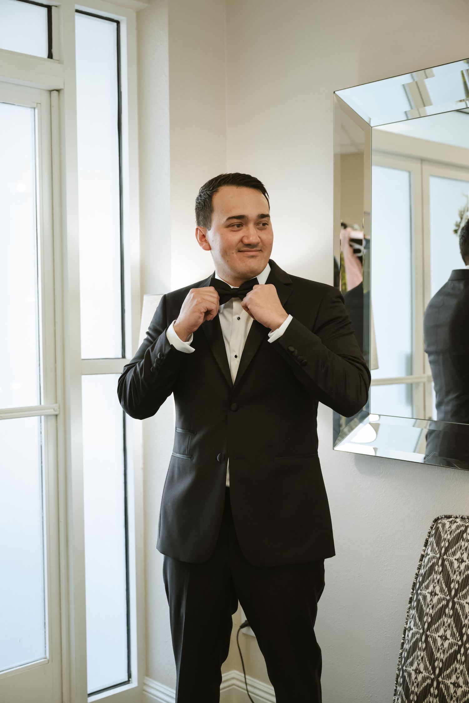 Groom in black tuxedo adjusting bow tie while smiling, standing near bright window with mirror reflection.