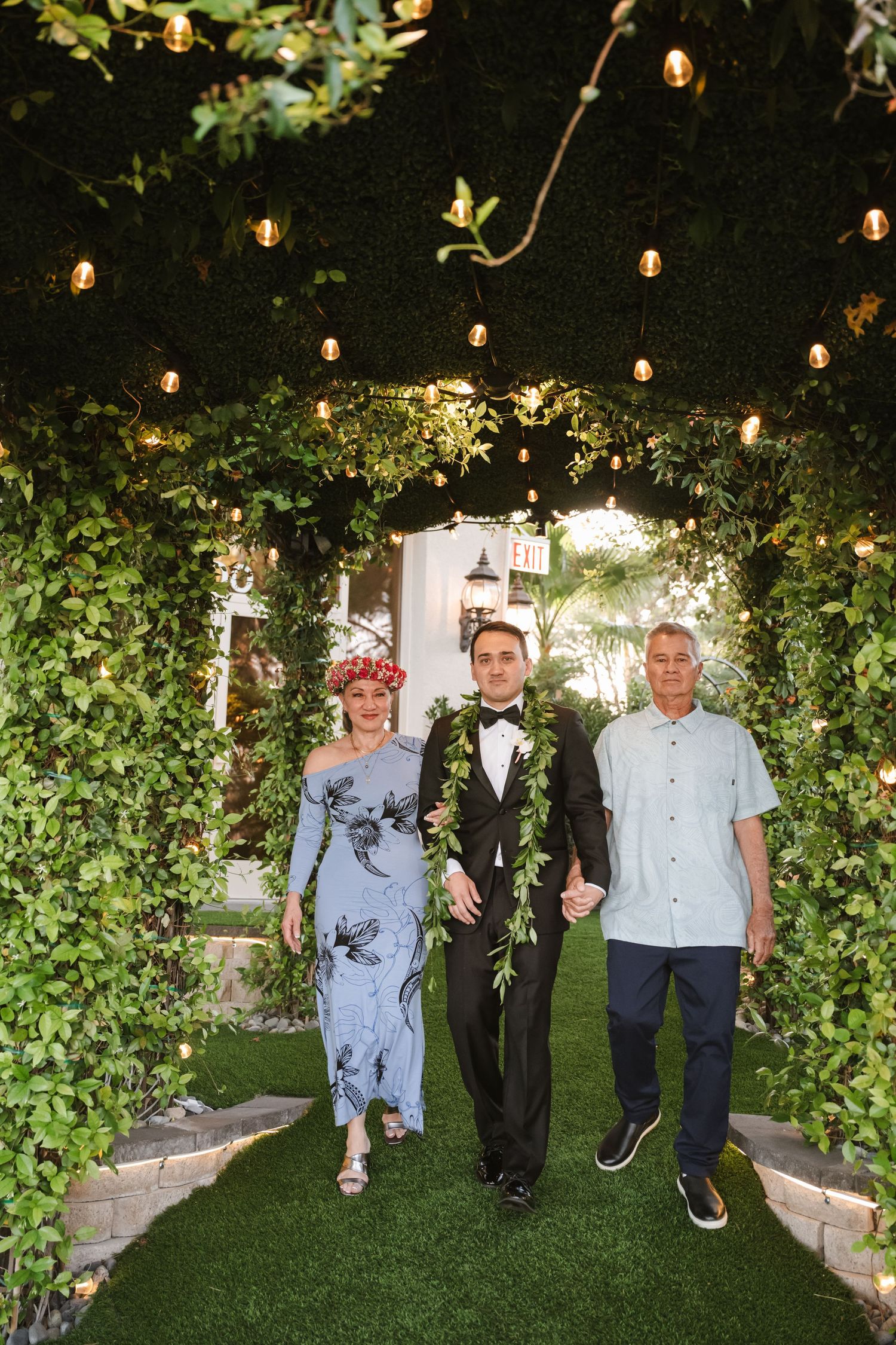 Groom in black tuxedo with green lei walks down garden aisle with parents under string lights.