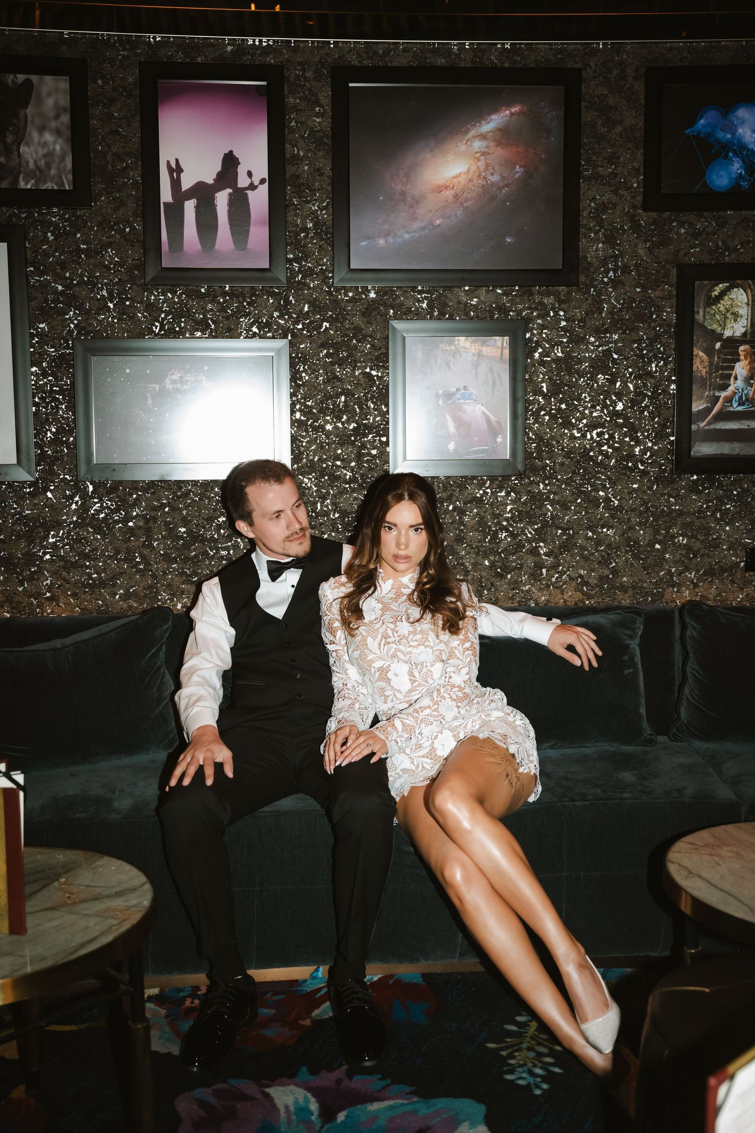 Elegant couple sitting on velvet sofa in upscale lounge, woman in lace dress, man in formal vest and bow tie.