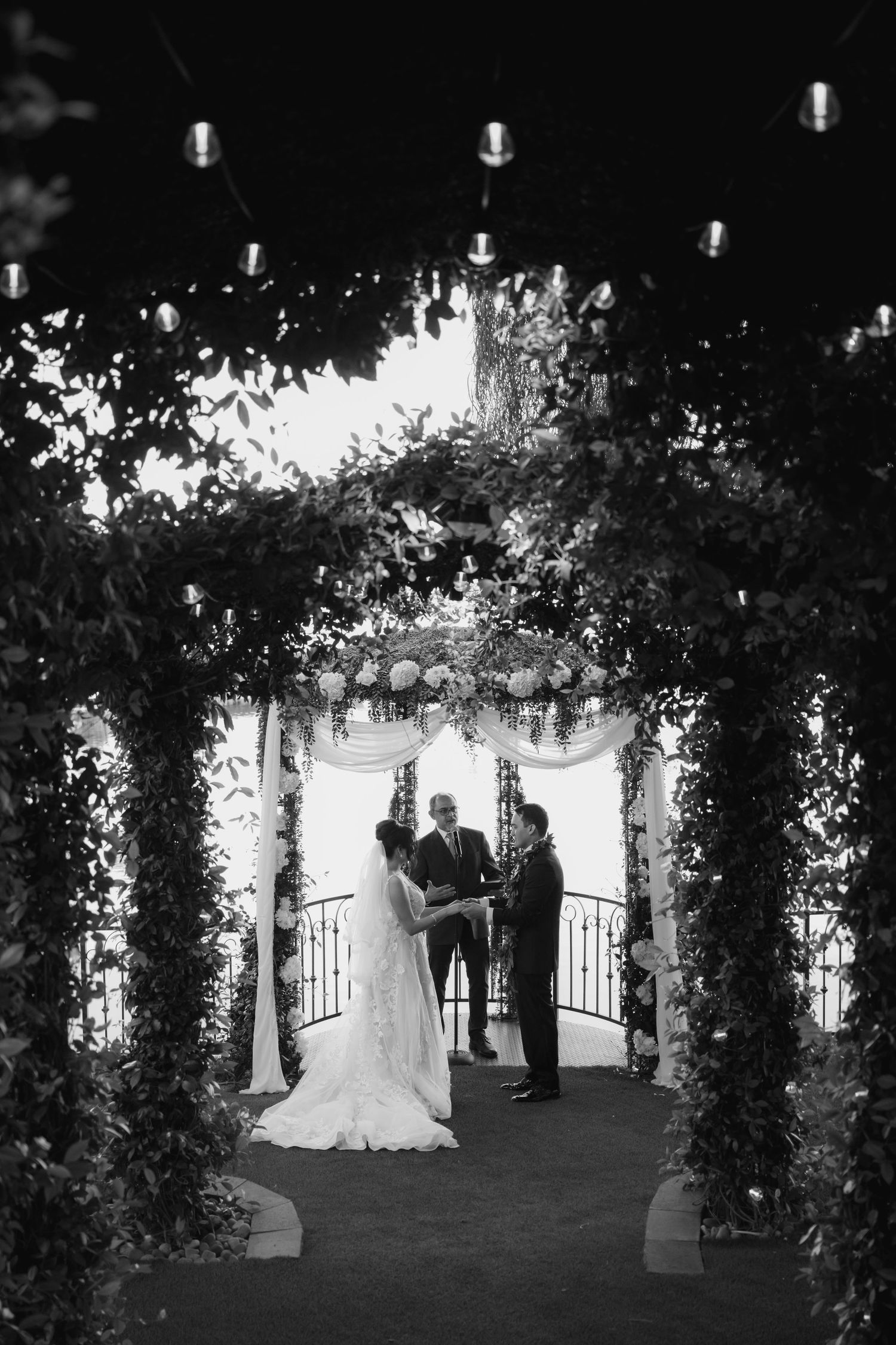 Black and white wedding ceremony photo under floral arch with string lights, bride and groom exchanging vows with officiant.