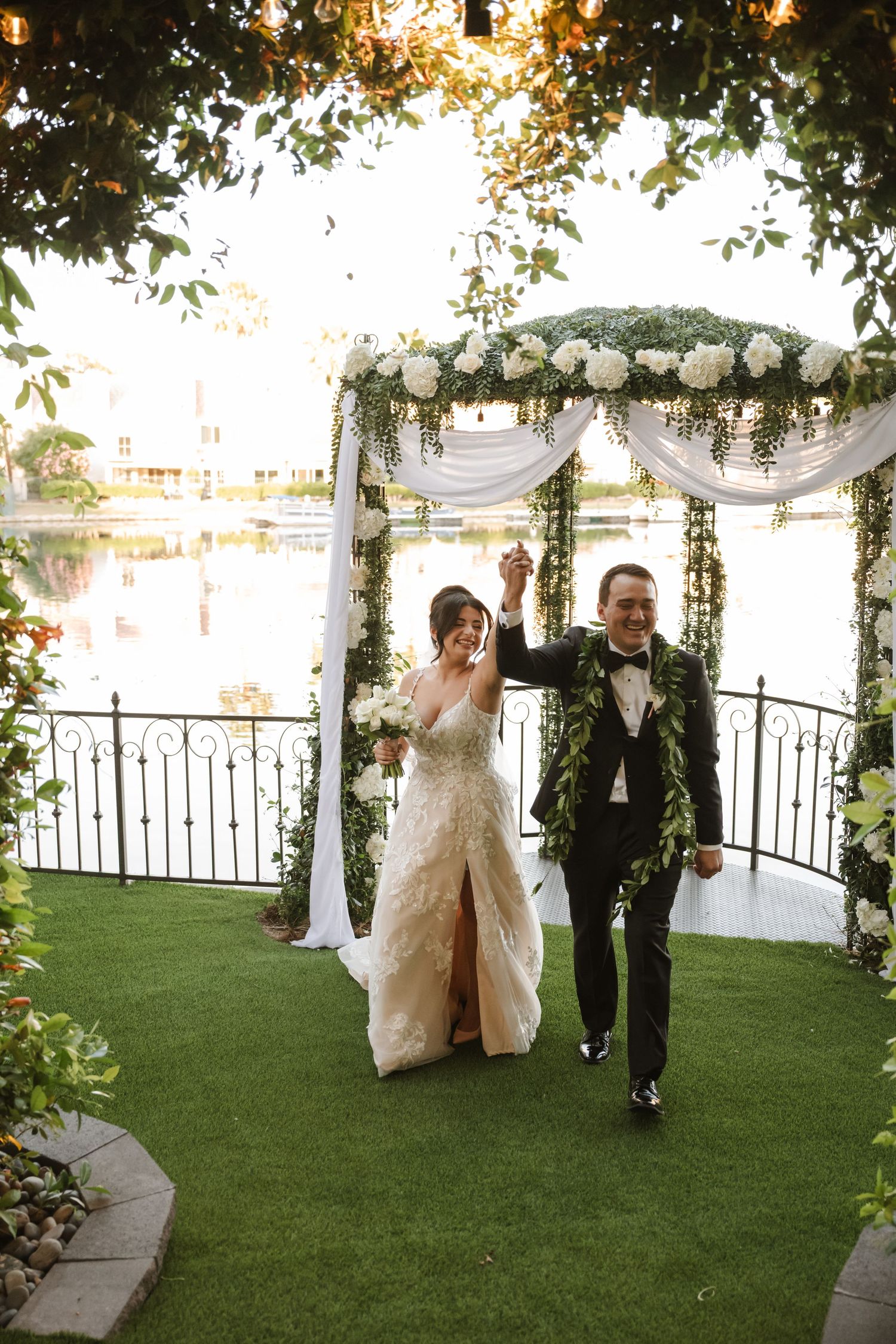 Newlyweds celebrate with raised hands under floral arch at waterfront outdoor wedding ceremony.