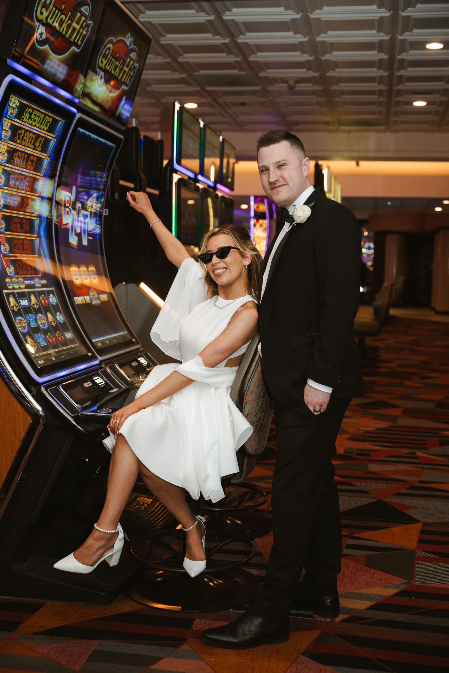 A bride in a short white dress plays slot machines while her groom stands beside her in a casino.