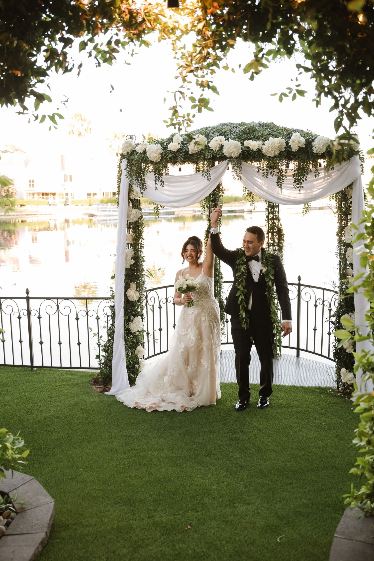 Bride and groom celebrate under a floral arch adorned with white flowers and greenery beside a scenic waterway.