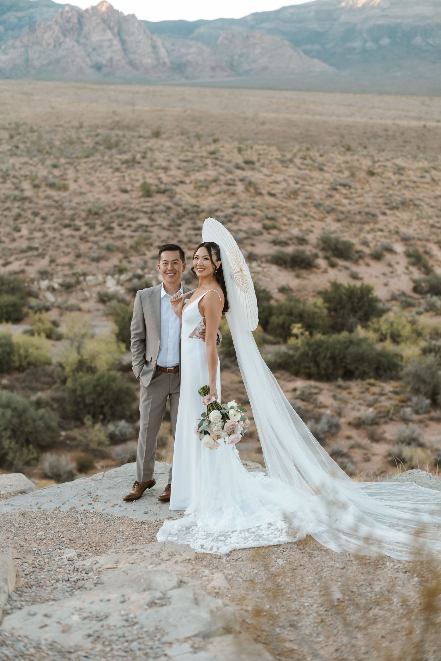 Bride and groom pose on desert rock at Red Rock Canyon, Nevada, with mountains and arid landscape backdrop.