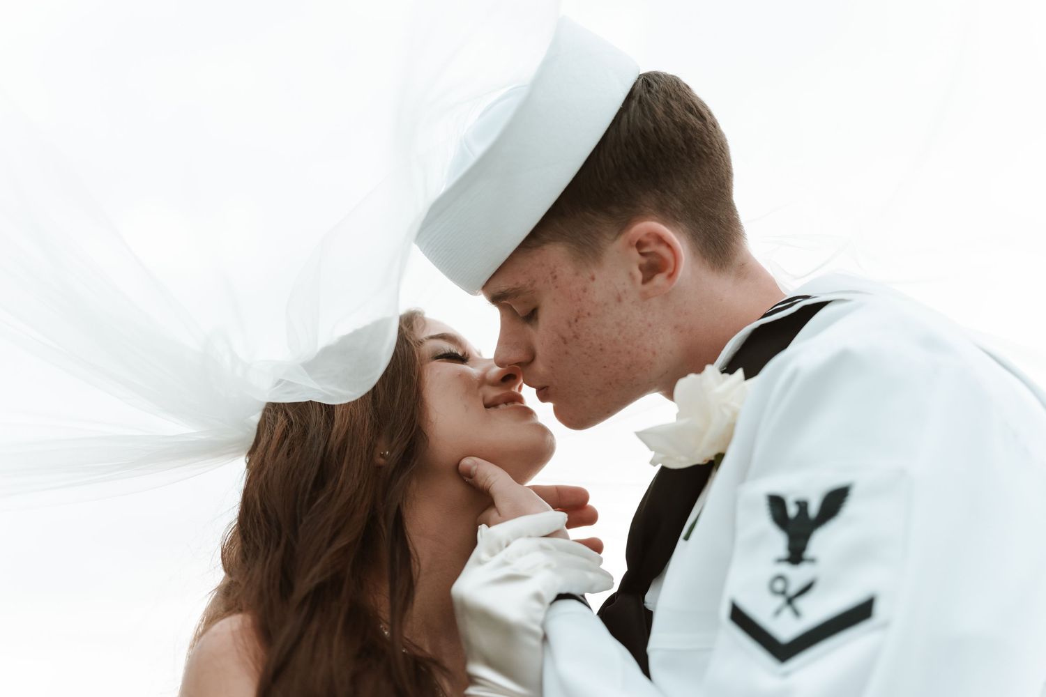 A Navy sailor in uniform shares a romantic kiss with his bride in a wedding veil, captured in a dreamy white-toned photo.