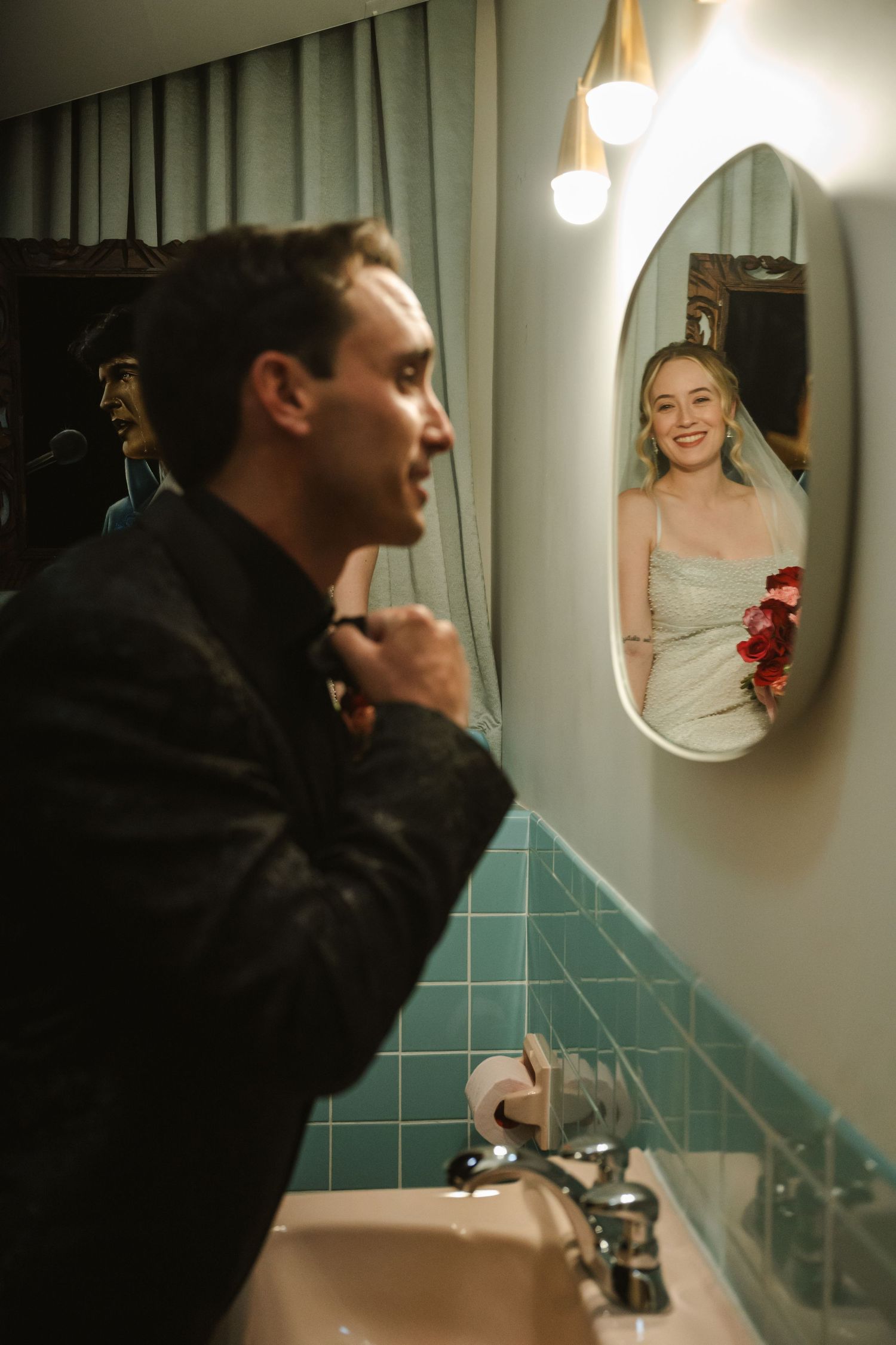 A groom adjusts his tie in a bathroom mirror reflecting a smiling bride in a white dress holding red roses.
