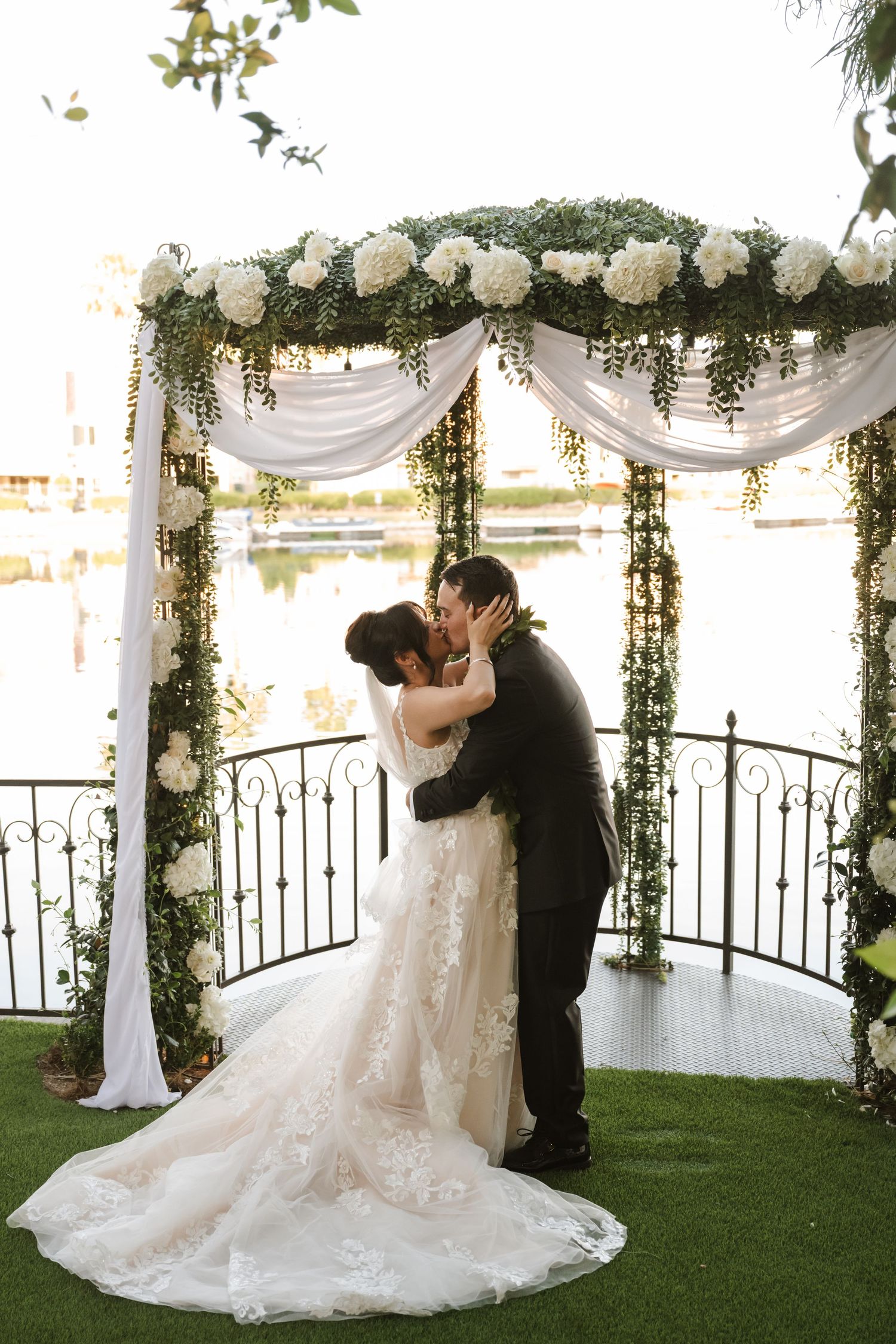Bride and groom share first kiss under white floral and greenery wedding arch overlooking lake.