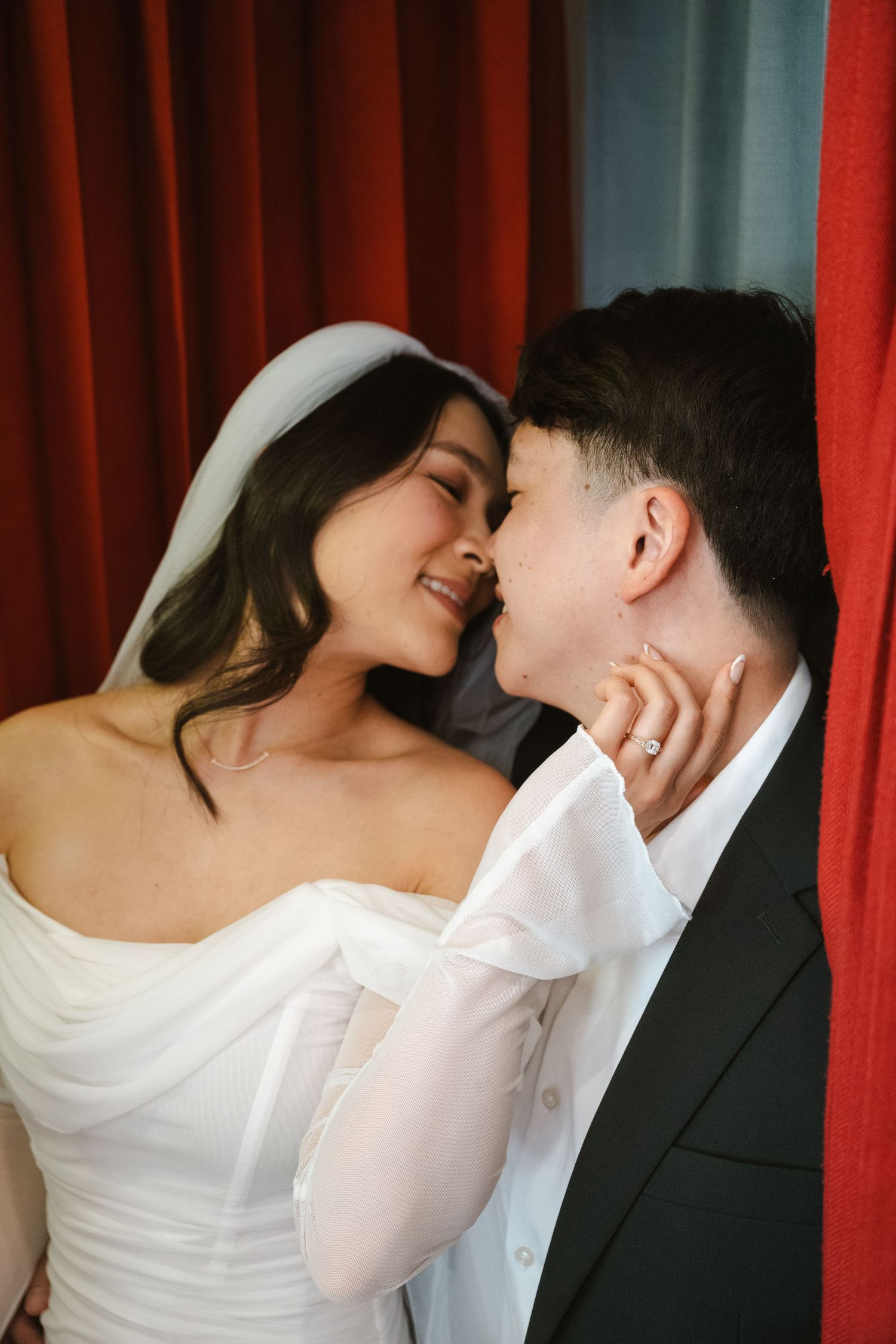 A bride in a white gown and veil shares a romantic moment with her partner in a suit near red curtains.