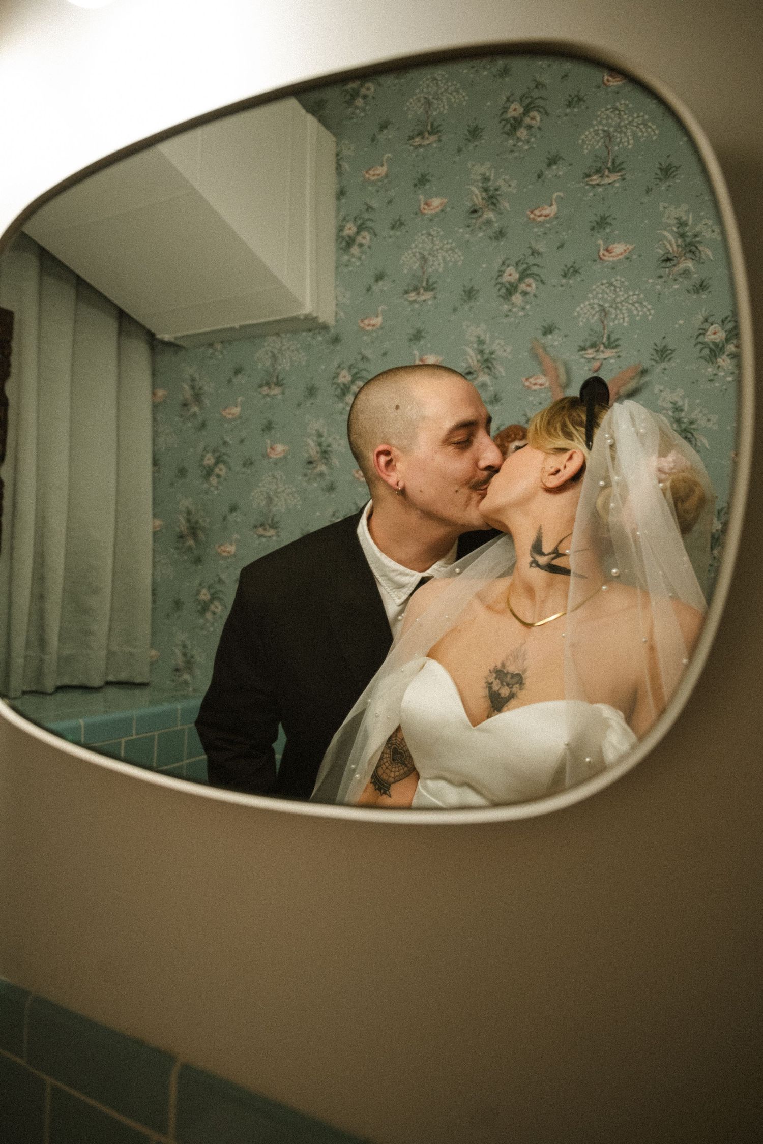 Tattooed bride and groom share a kiss reflected in a vintage bathroom mirror, bride wearing white dress and veil.