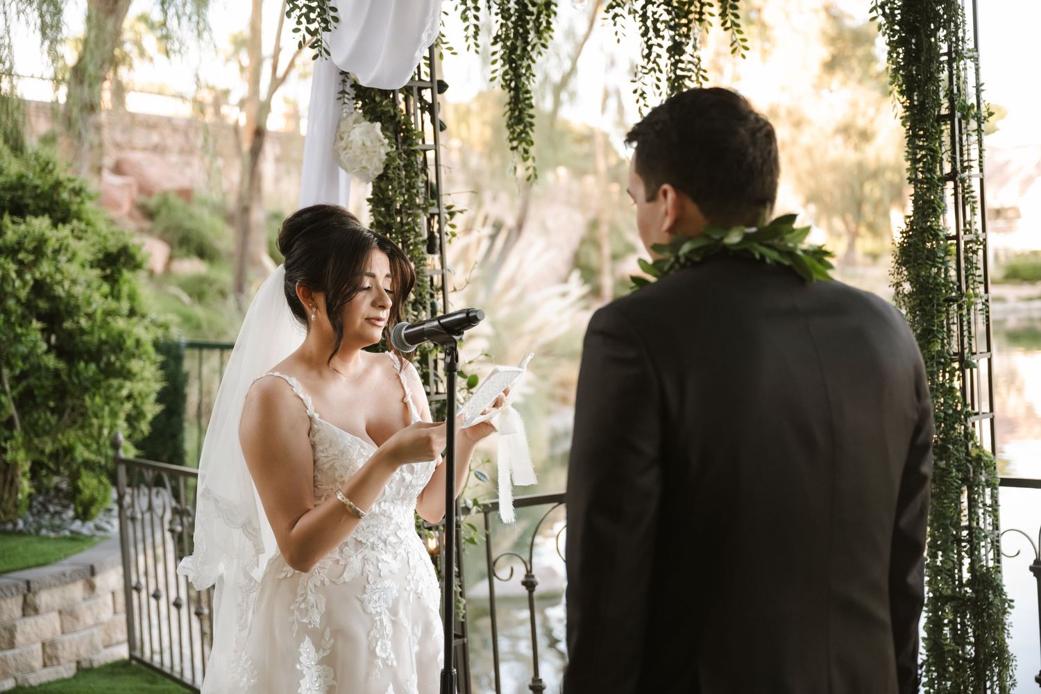 Bride in lace gown reads wedding vows to groom wearing floral lei at elegant outdoor ceremony.