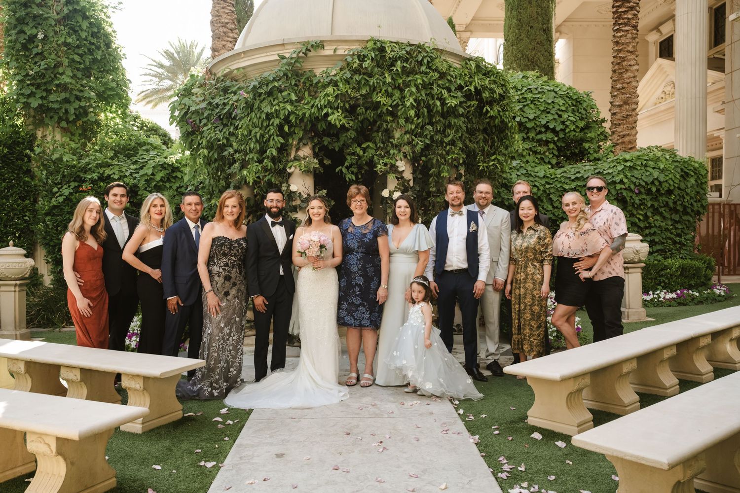 Wedding party gathers outdoors in a garden setting with palm trees and stone benches.