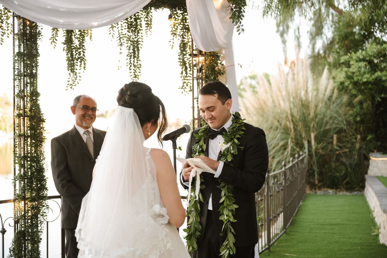 Groom in black tuxedo with green lei exchanges vows with bride in white gown at outdoor wedding ceremony.