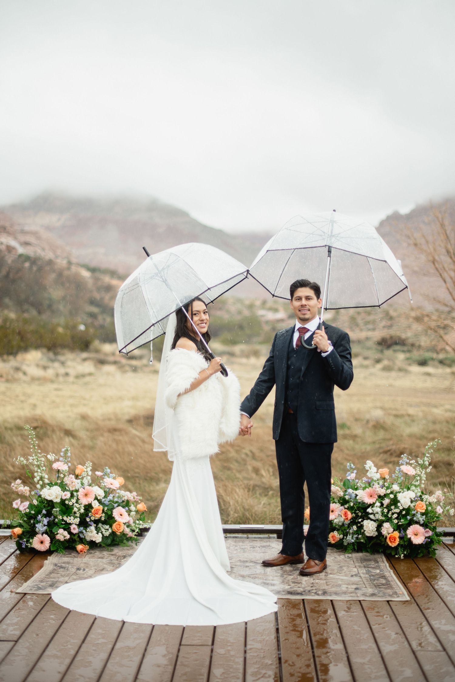 Bride and groom holding clear bubble umbrellas on a rainy wedding day, standing on a wooden deck with floral arrangements.