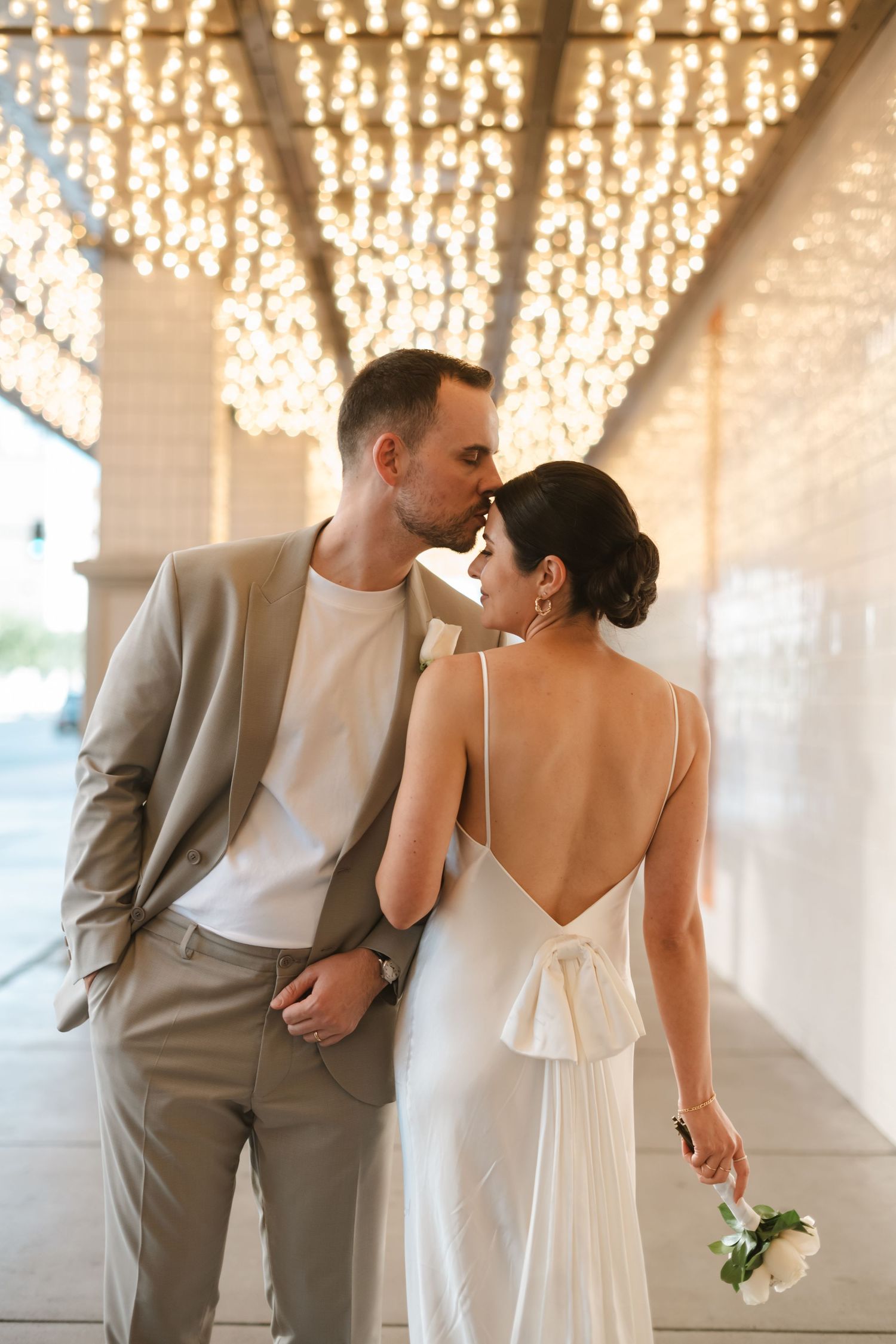 Romantic couple in wedding attire embracing under a canopy of glowing string lights at an elegant venue.