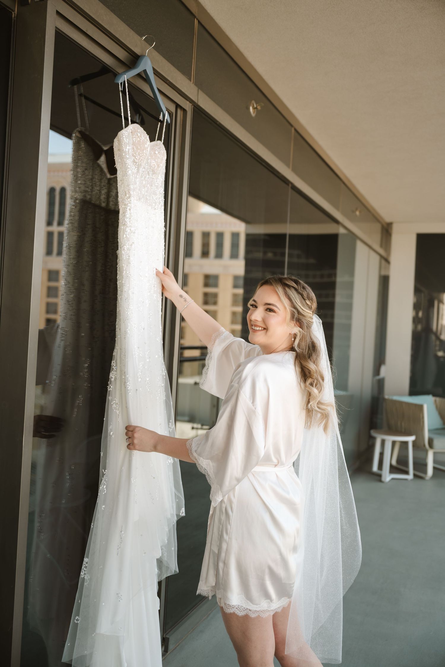 A white wedding dress hangs on display against glass windows in a modern building.