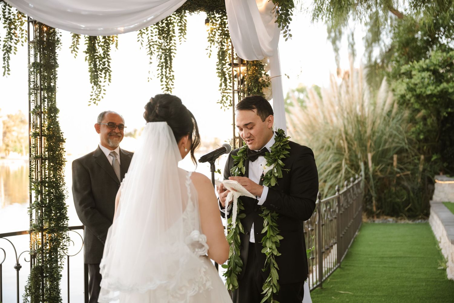 Groom in black suit with lei placing ring on brides finger during outdoor wedding ceremony.