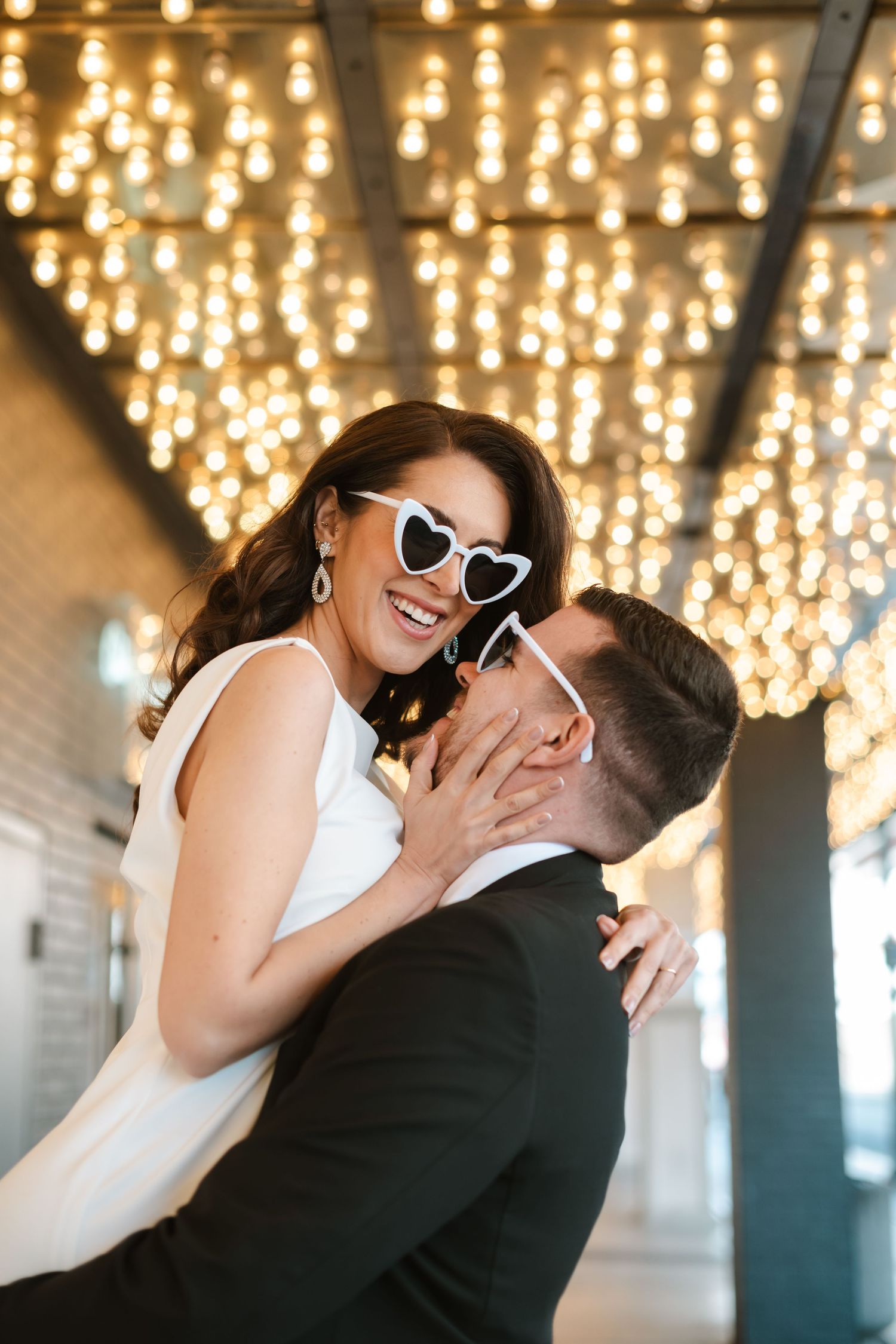 A couple wearing sunglasses poses playfully under twinkling ceiling lights.
