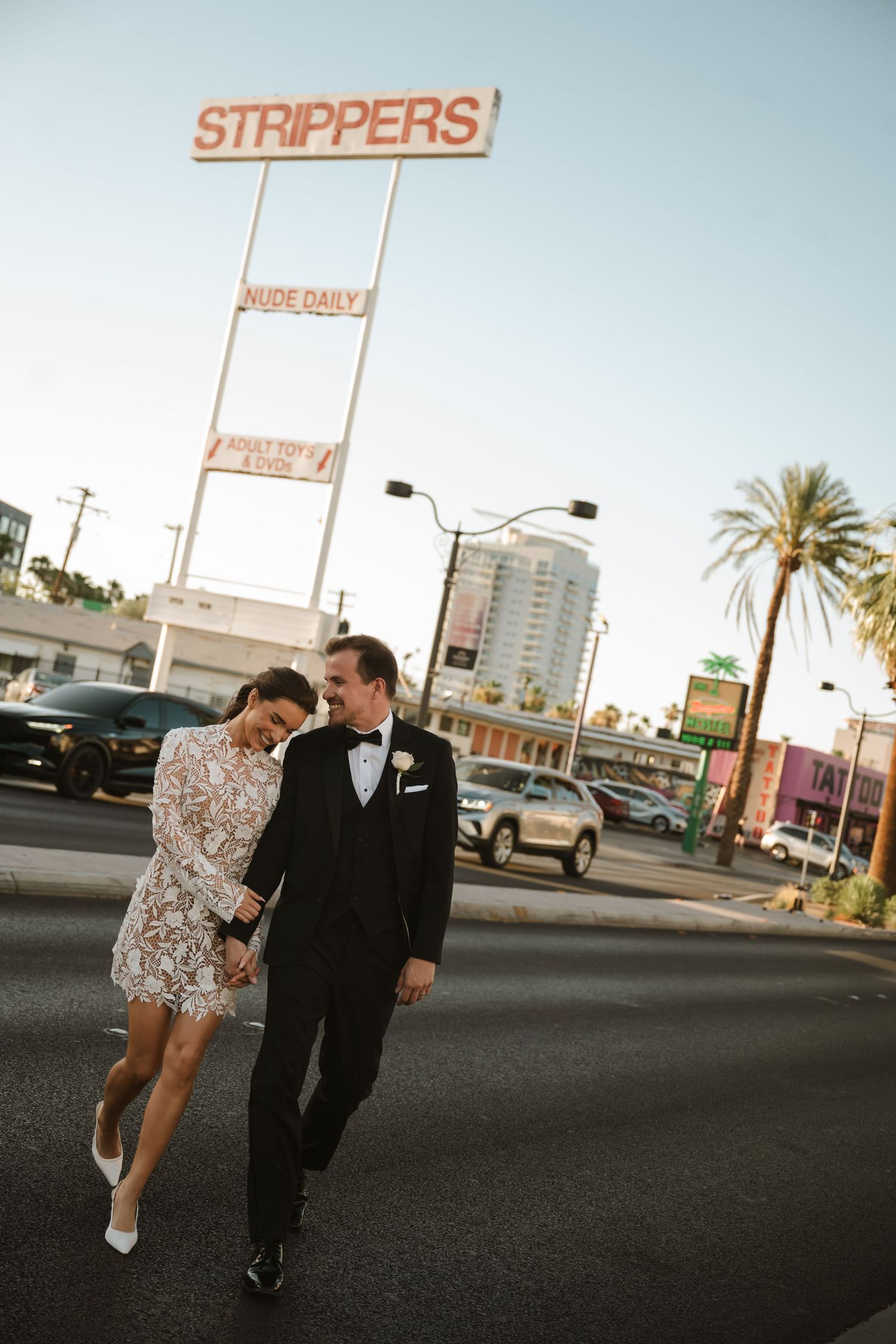 Newlywed couple in wedding attire walking hand in hand on Las Vegas Strip street with neon signs in background.