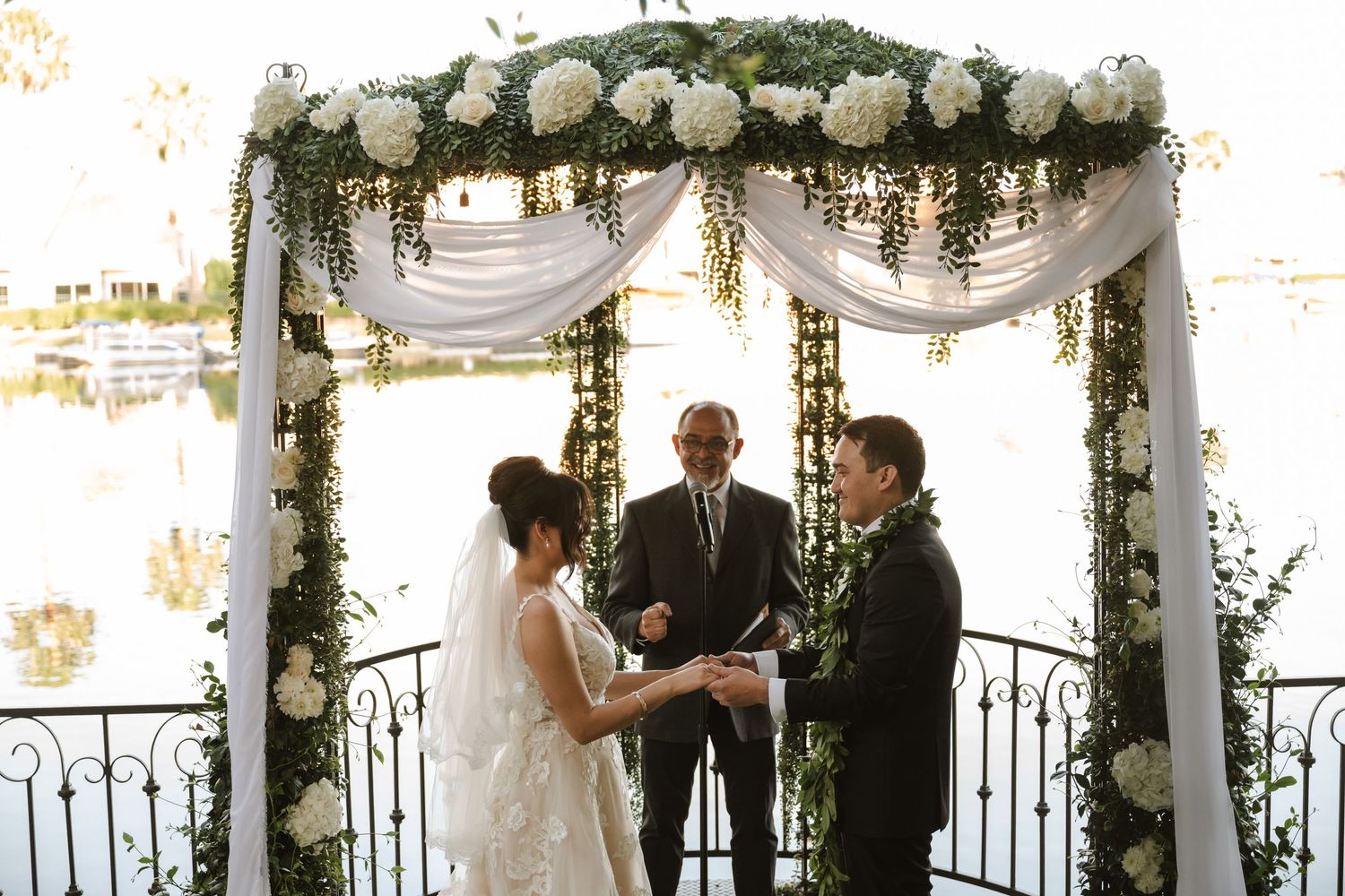Bride and groom exchange vows under a floral arch adorned with white roses and greenery by a lakeside.