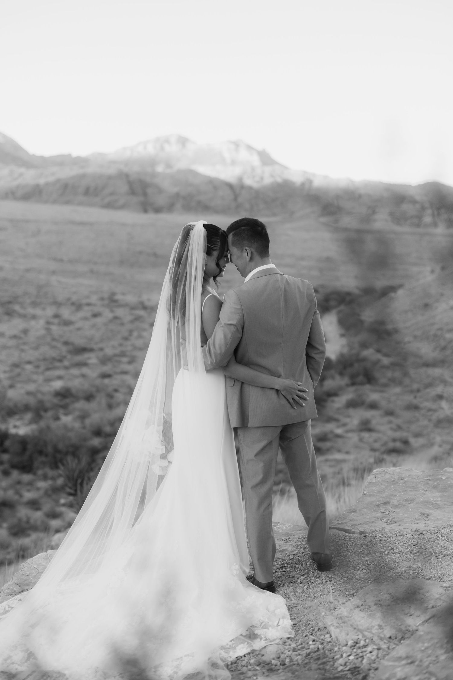 Black and white photo of bride and groom embracing with mountain landscape backdrop on their wedding day.