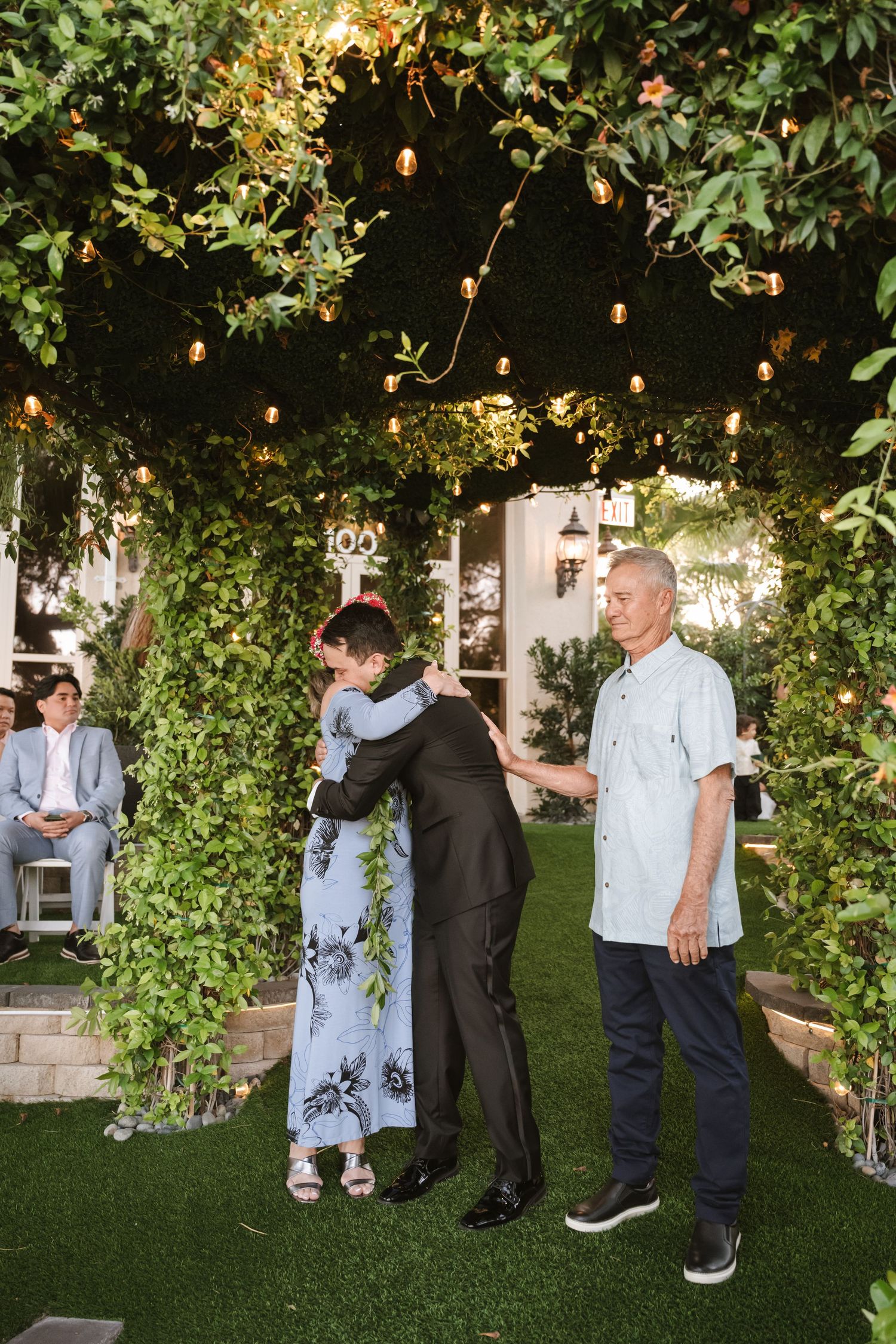 Outdoor wedding ceremony with guests hugging under string lights and lush green ivy-covered walls on a grass lawn.