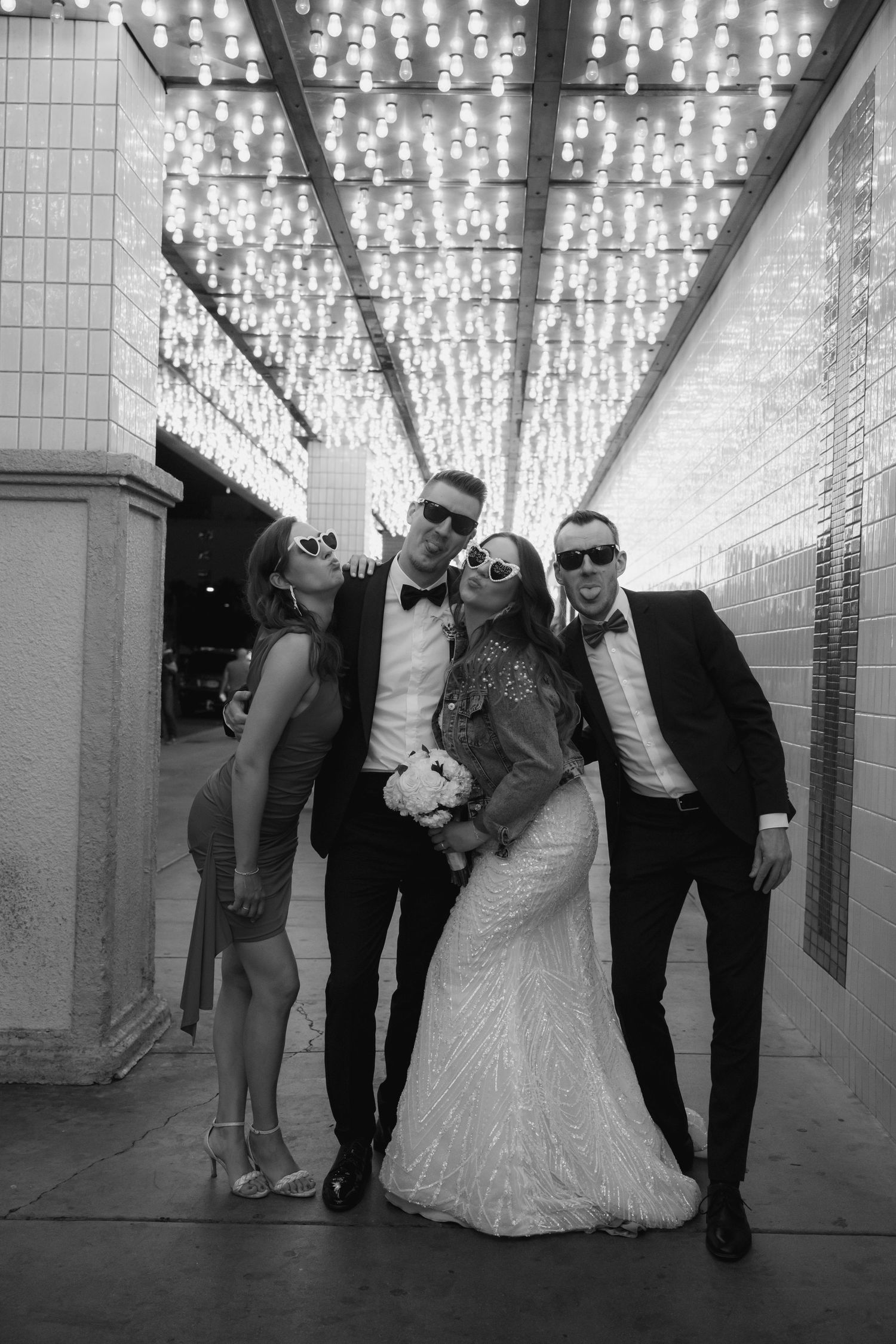 Wedding group strikes fun poses under ceiling of string lights in black and white.