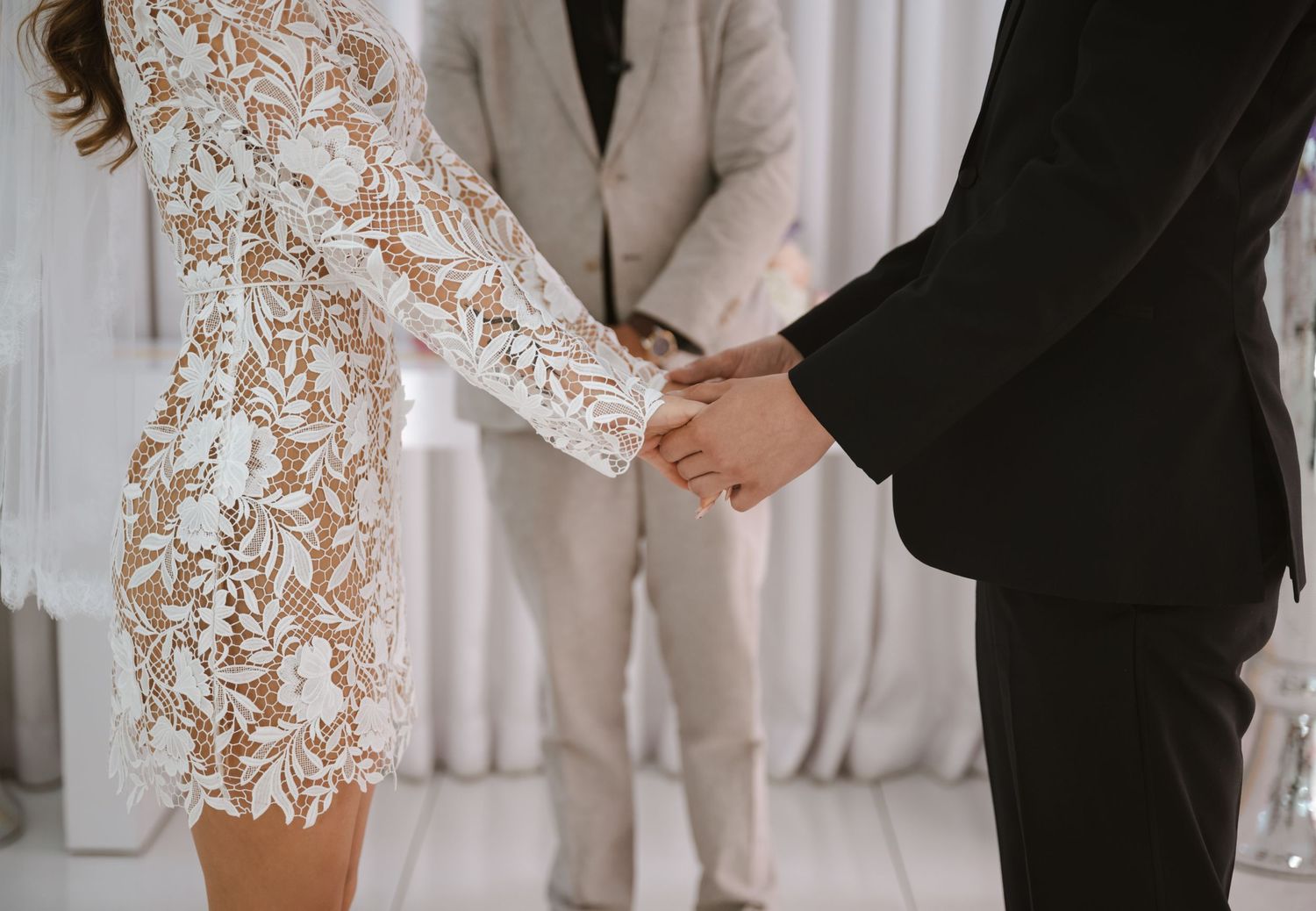 Couple holding hands in wedding attire, bride in lace dress, officiant in background at elegant ceremony.