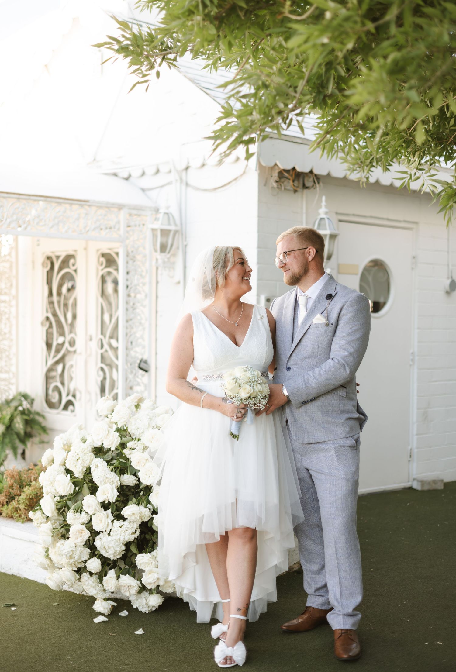 A couple in wedding attire poses together against a white building with floral decorations.