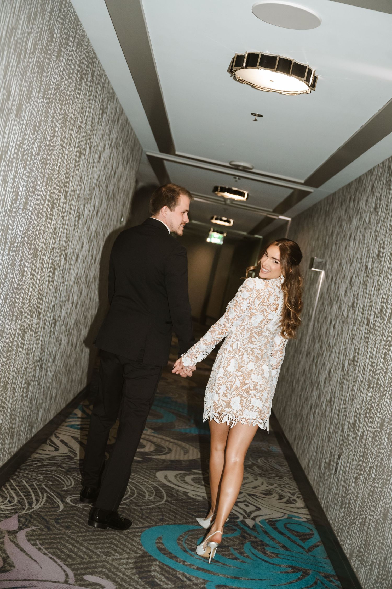 Couple in formal attire holding hands walking down a hotel corridor, woman in lace mini dress and heels.