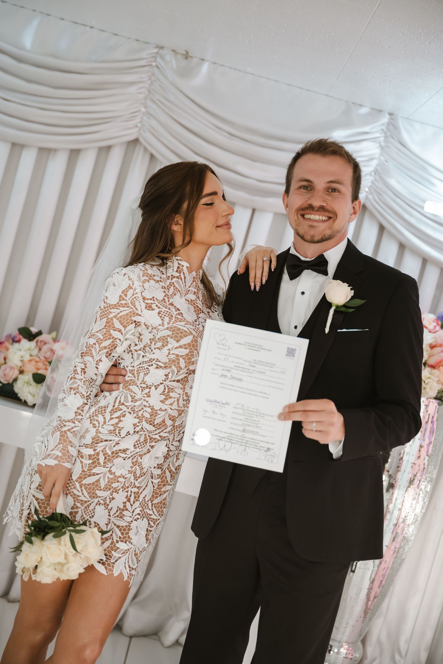 Newlyweds smiling and holding marriage certificate in decorated wedding venue.