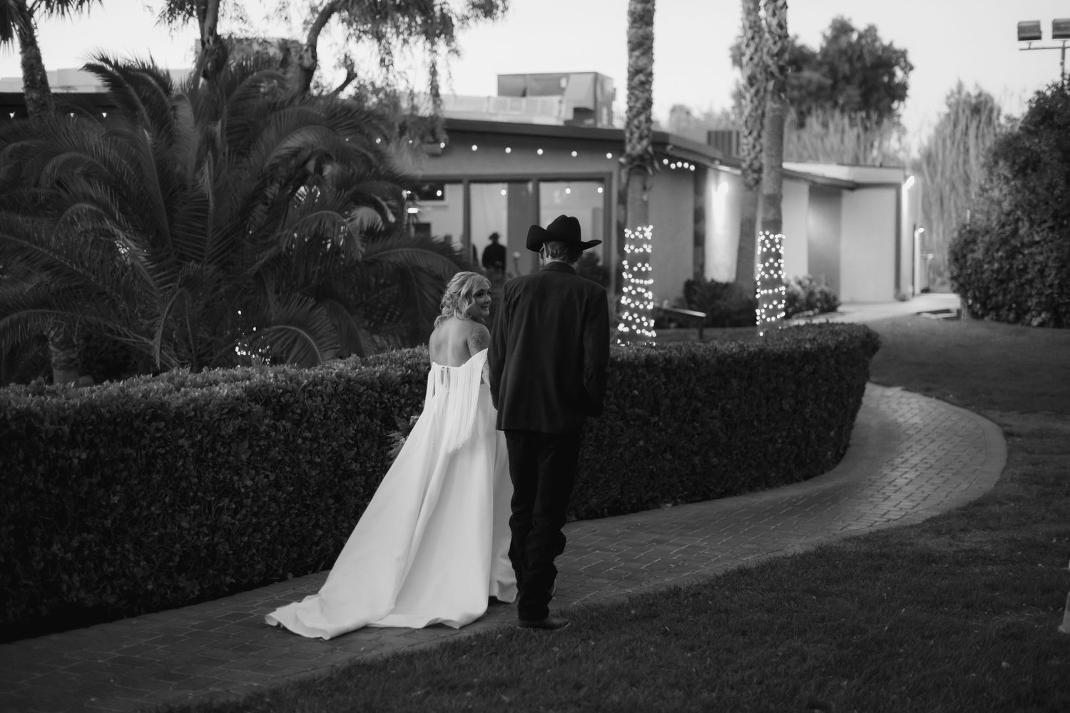 Black and white photo of a bride and groom walking hand in hand toward a building surrounded by lush greenery.